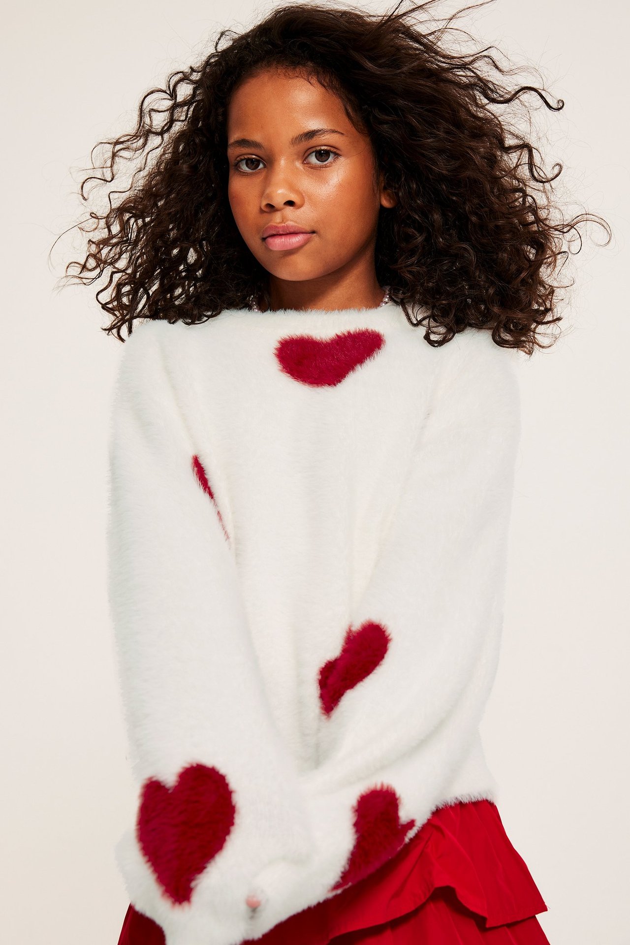 Framifrån: ett barn i en offwhite fluffig tröja med röda hjärtan och en röd kjol.