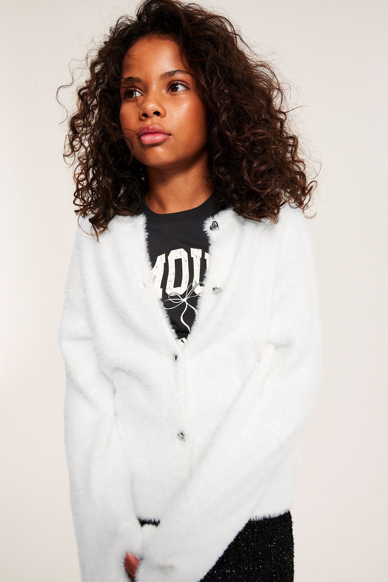 Forfra, et barn i en offwhite, fluffy cardigan med hjerteknapper over en svart trykt t-skjorte.