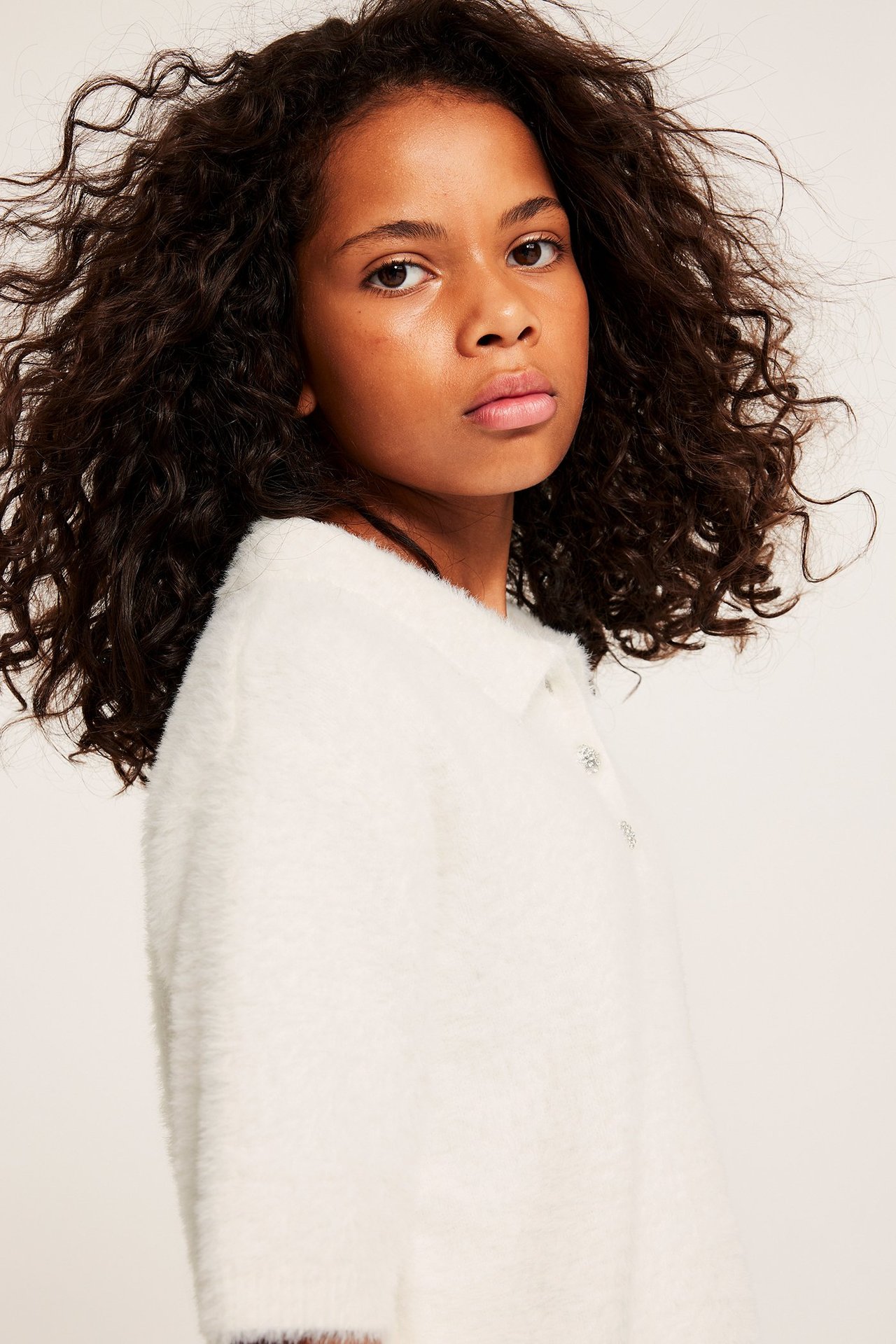 Närbild, sidovy: Barn bär en offwhite stickad tröja i fjädergarn med kort ärm, krage, knappar.