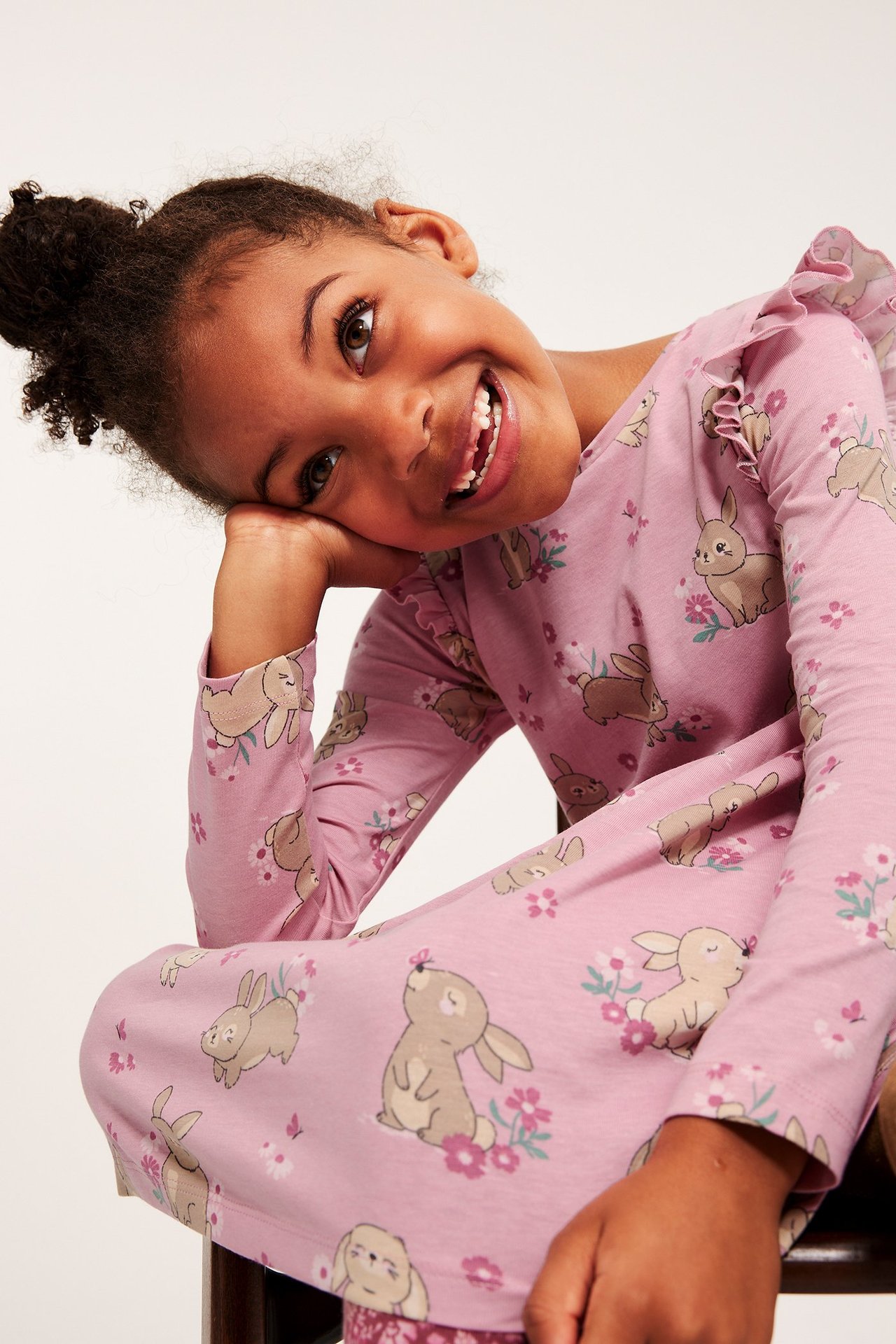 Nærbilde av et smilende barn i rosa bomullskjole med kaninmønster og volanger på skuldrene.