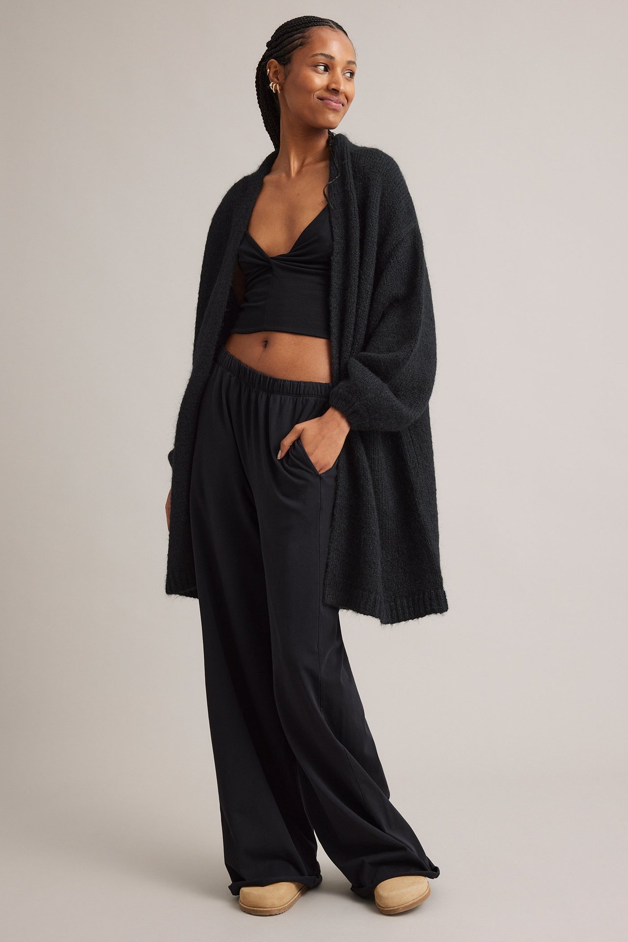 Edestä, nainen mustassa avoimessa villasekoitekardigaanissa, mustassa crop topissa, leveälahkeisissa housuissa ja puukengissä.