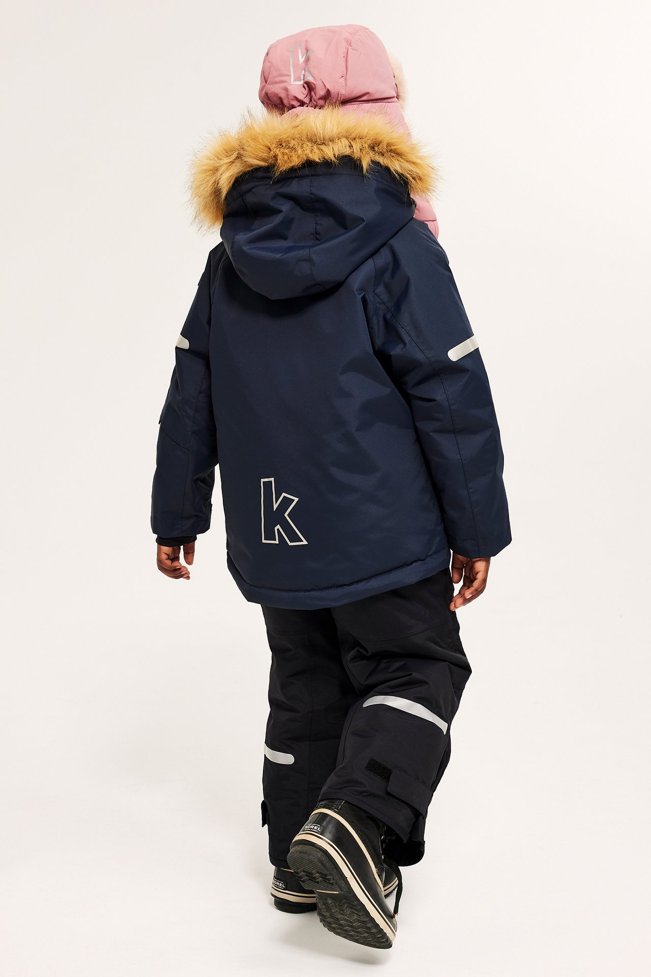 Widok z tyłu dziecka w granatowej kurtce zimowej z futrzanym kapturem, czarnych spodniach narciarskich i czarnych butach.