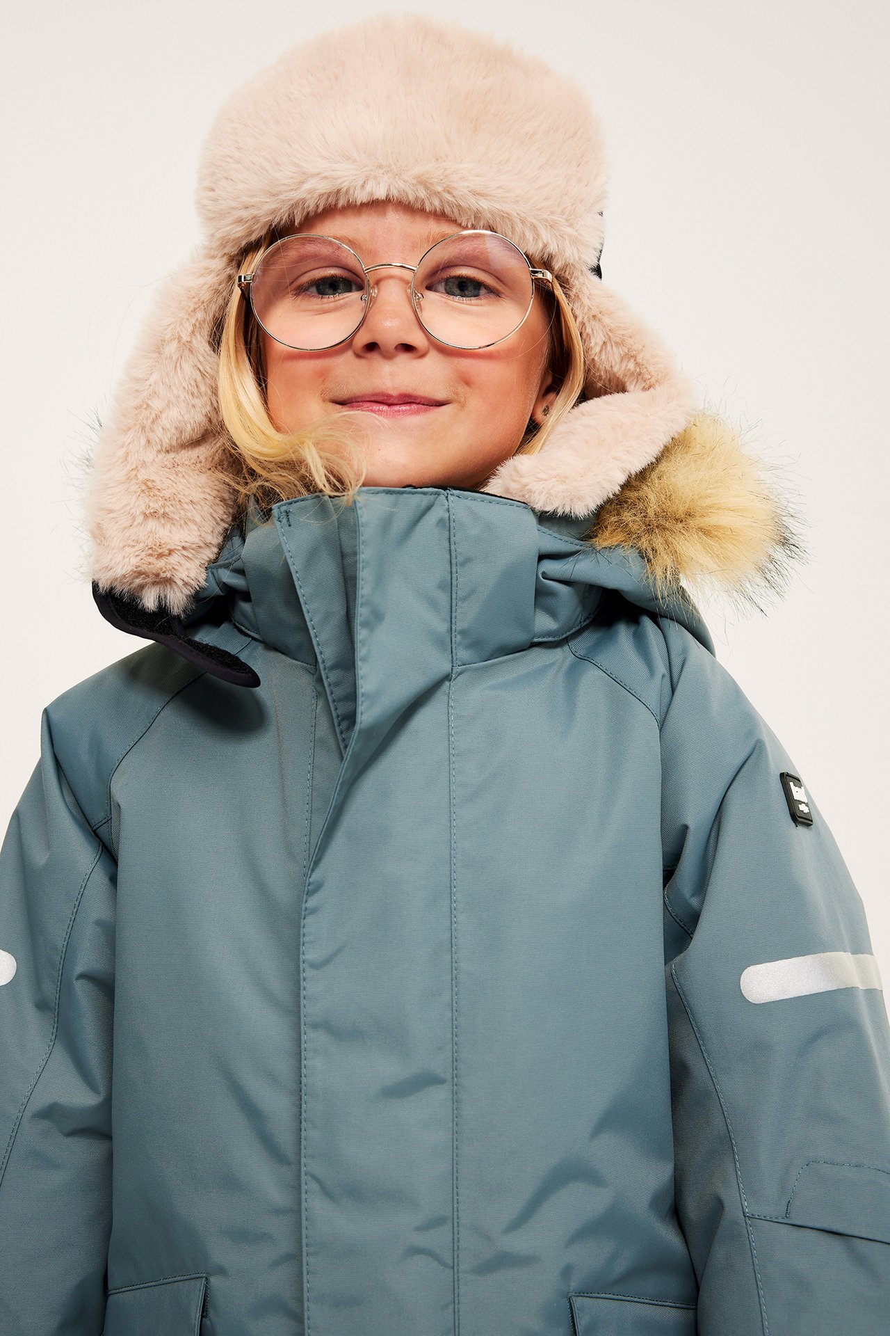 Närbild framifrån på ett barn i blå vinterjacka med pälskantad huva, beige fuskpäls mössa och runda glasögon.