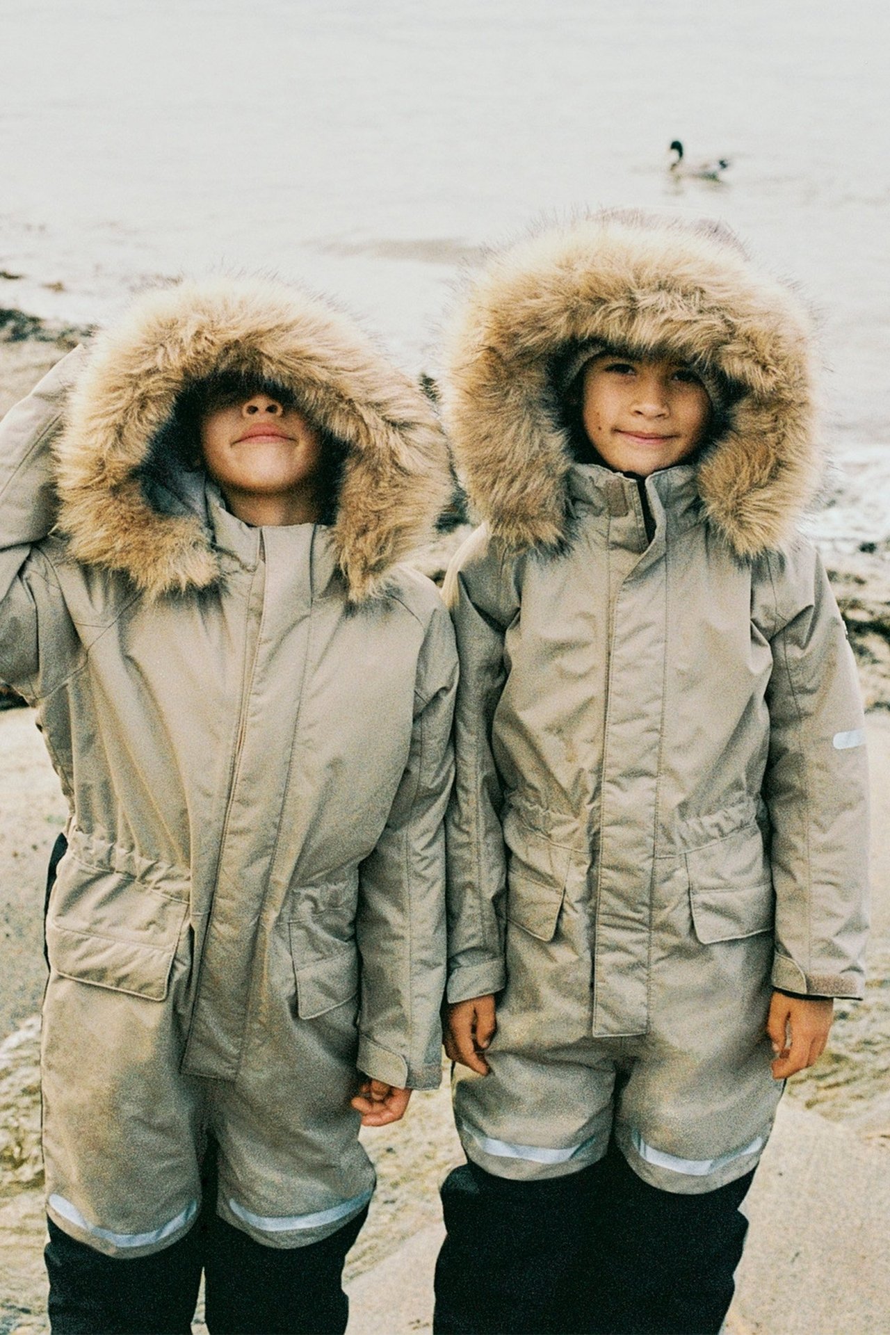 Widok z przodu dwóch dzieci w beżowych kombinezonach zimowych z obszytymi futrem kapturami i odblaskami.
