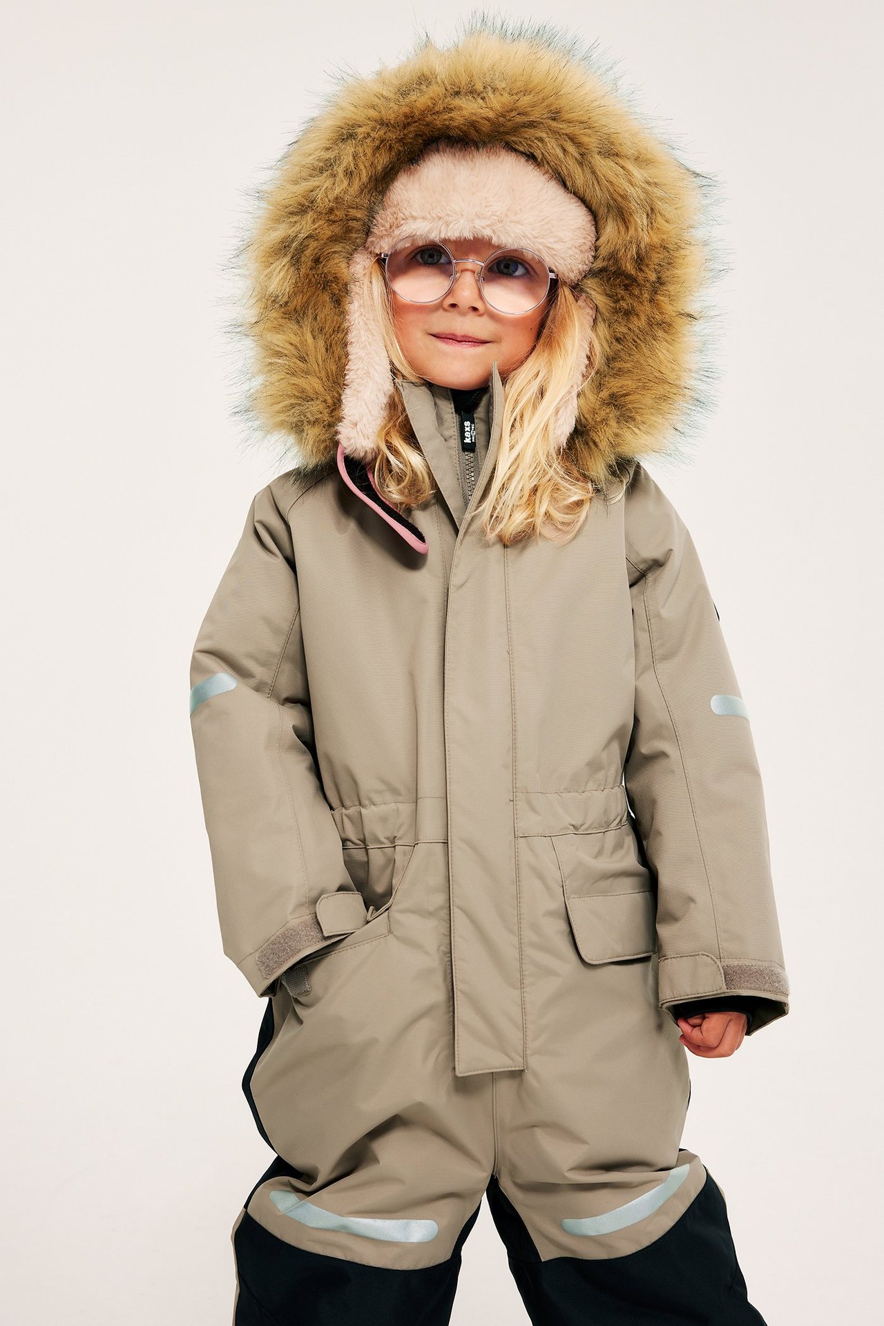 Widok z przodu dziecka w beżowym kombinezonie zimowym z kapturem obszytym futrem, różowej czapce i okrągłych okularach.