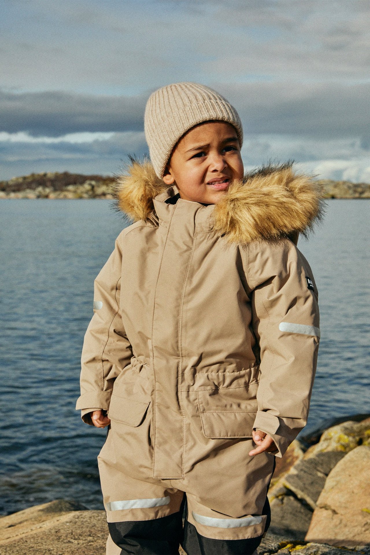 Edestä: Lapsi beigessä talvihaalarissa tekoturkishupulla, heijastimilla ja beigessä neulotussa pipossa.