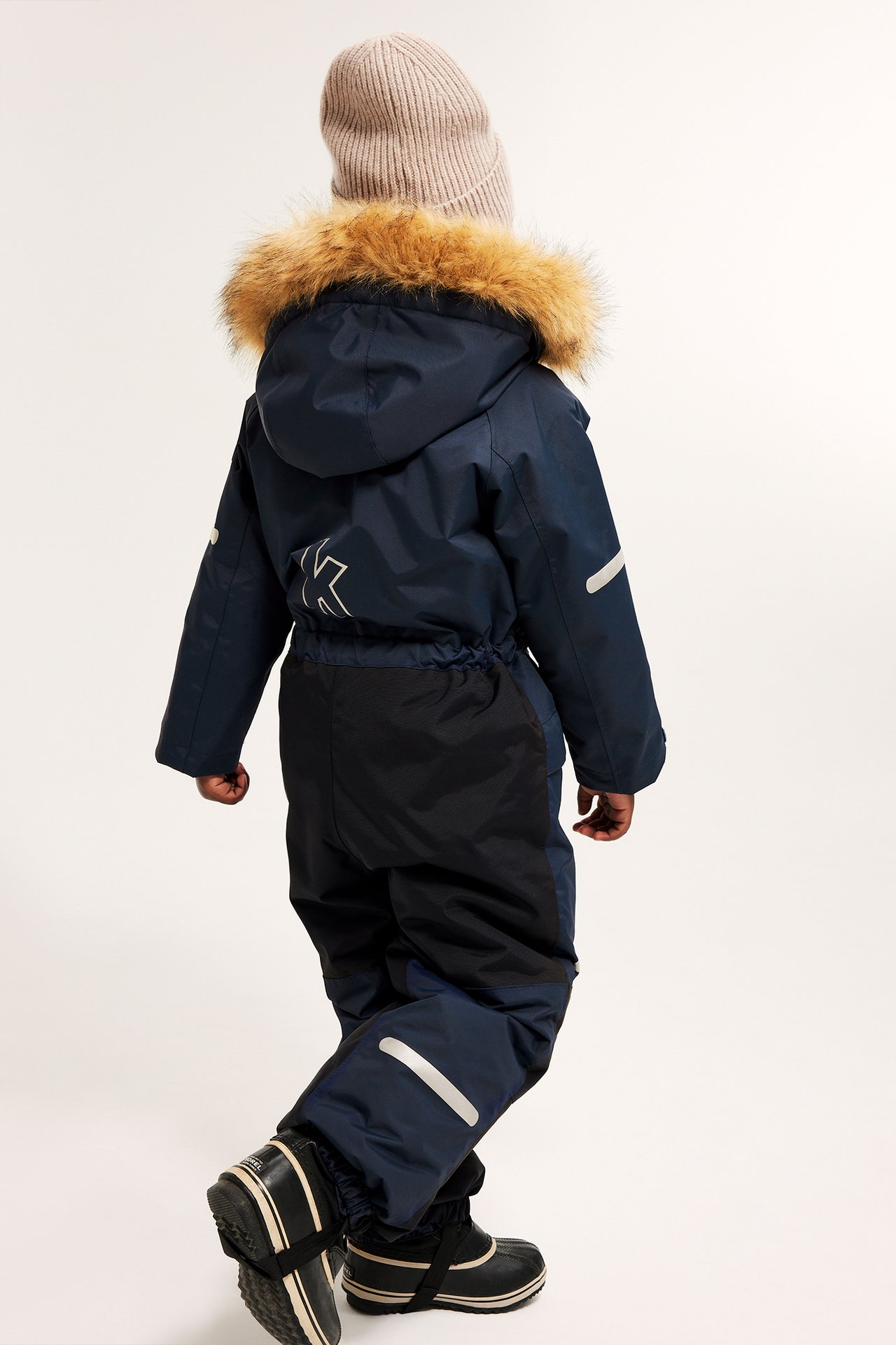 Widok z tyłu dziecka w granatowym kombinezonie zimowym z kapturem obszytym futrem, beżową czapką i czarnymi butami.