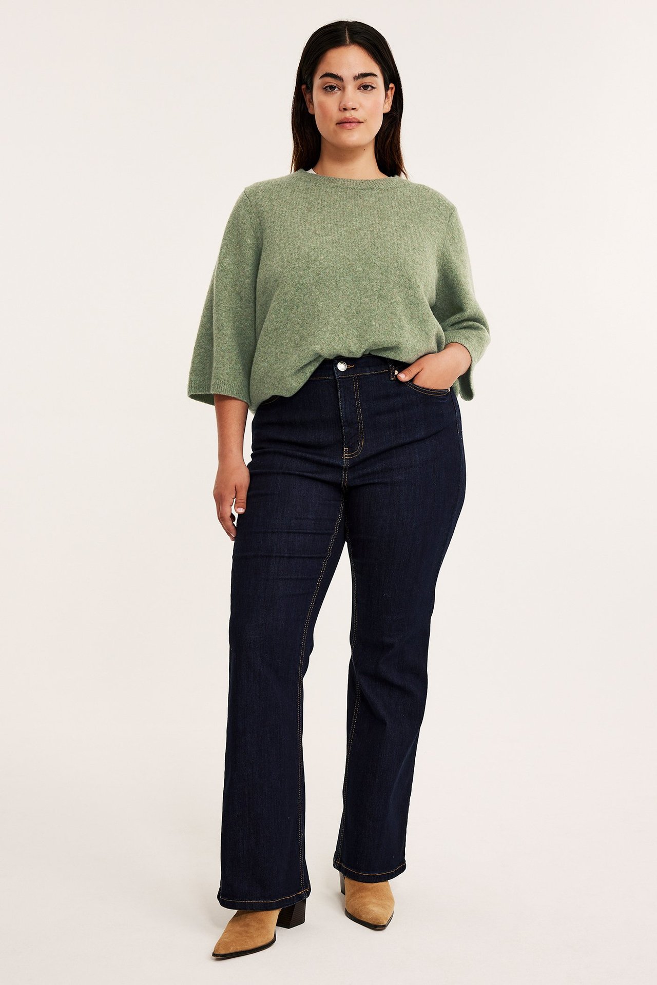 Edestä: nainen yllään väljä meleerattu vihreä neuletoppi 3/4-hihoilla, tummansiniset farkut ja beiget korkokengät.