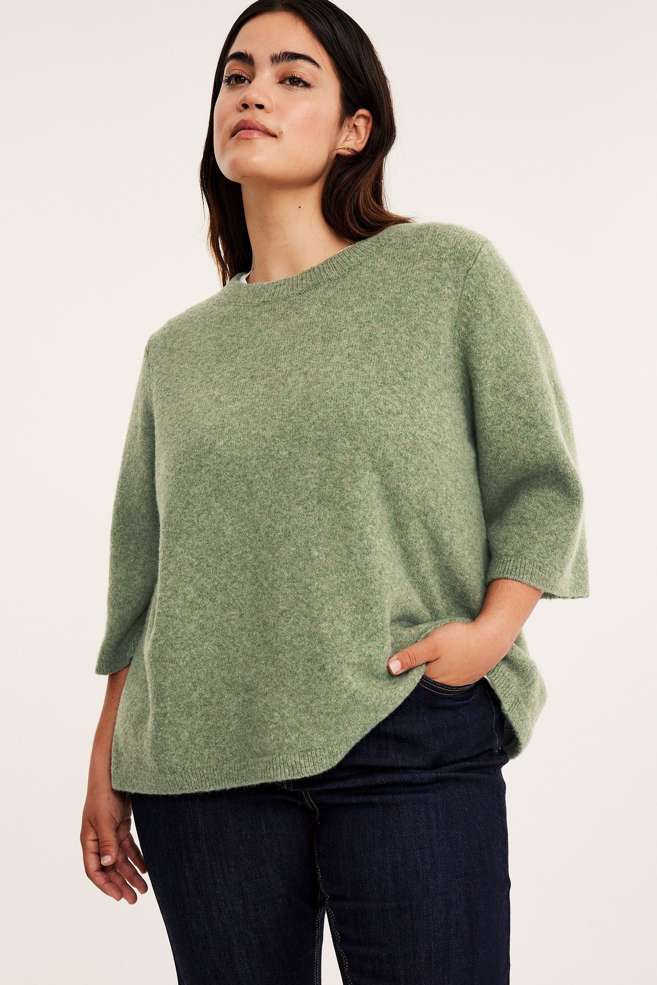 Edestä, nainen yllään vihreä meleerattu neuletoppi 3/4-hihoilla ja tummansiniset farkut.