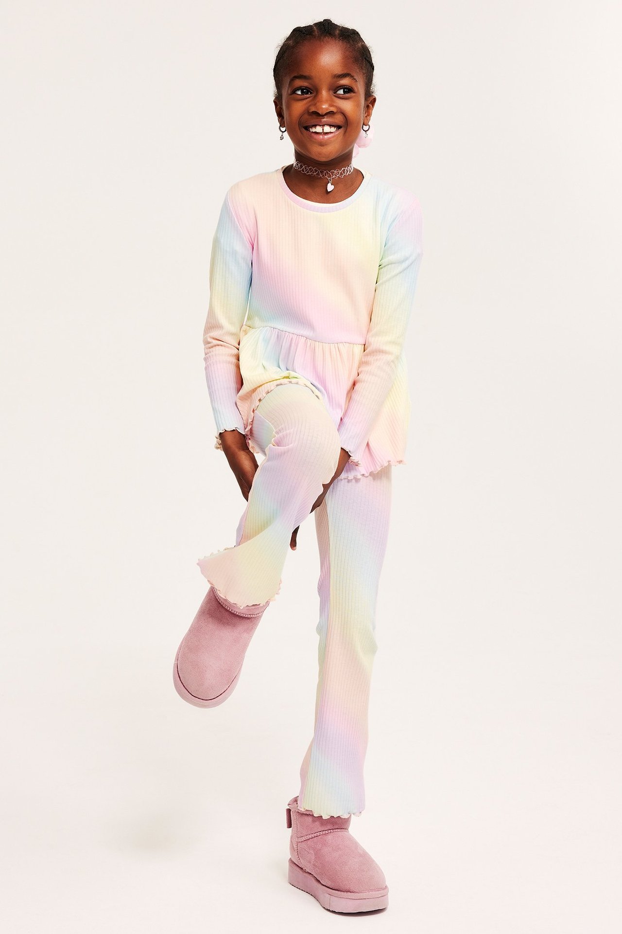 Widok z przodu: Dziecko w tęczowych prążkowanych legginsach flare, pasującym topie i różowych butach.
