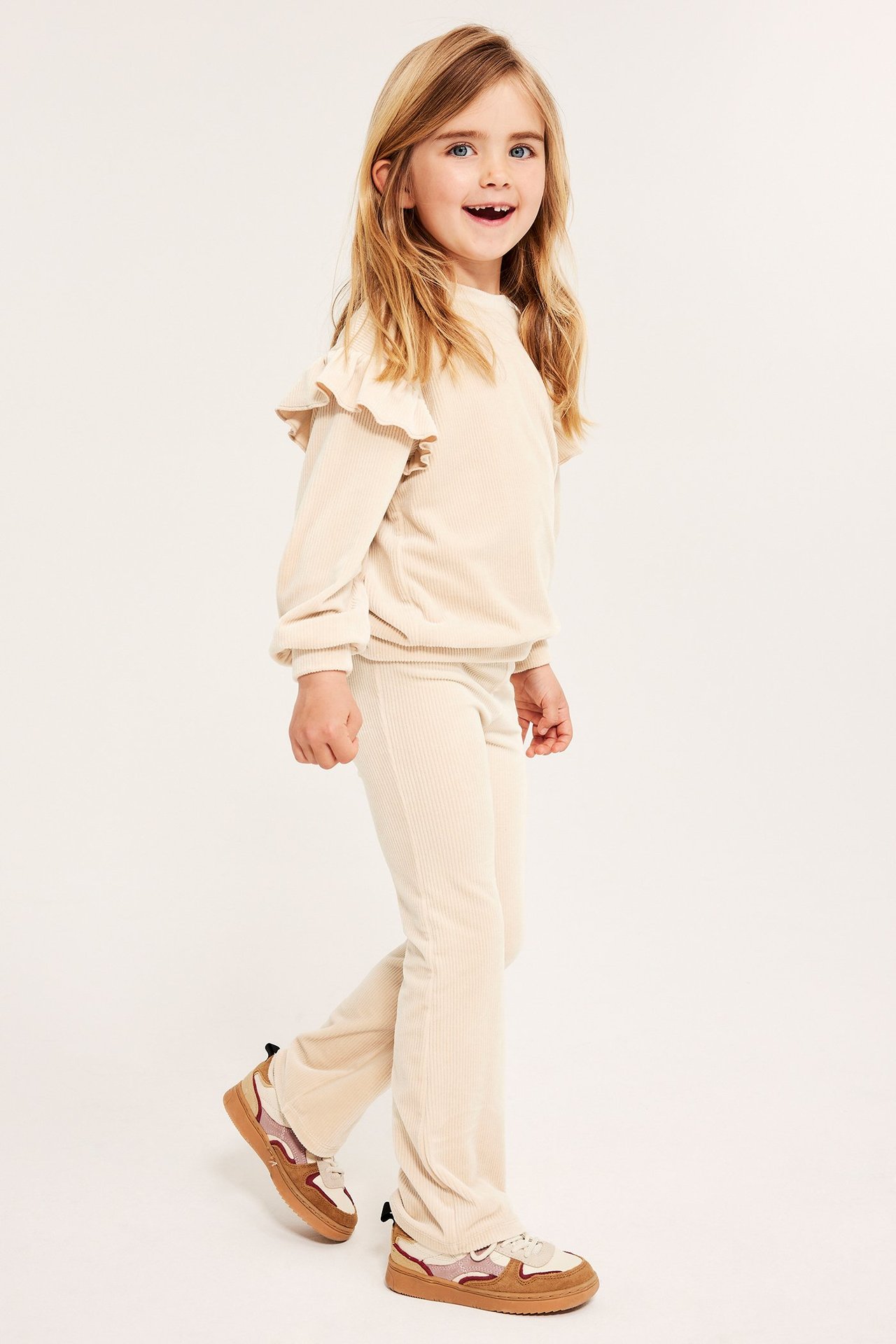 Sidevisning av et barn i beige ribbet velour-genser med volanger, matchende bukser og flerfargede joggesko.