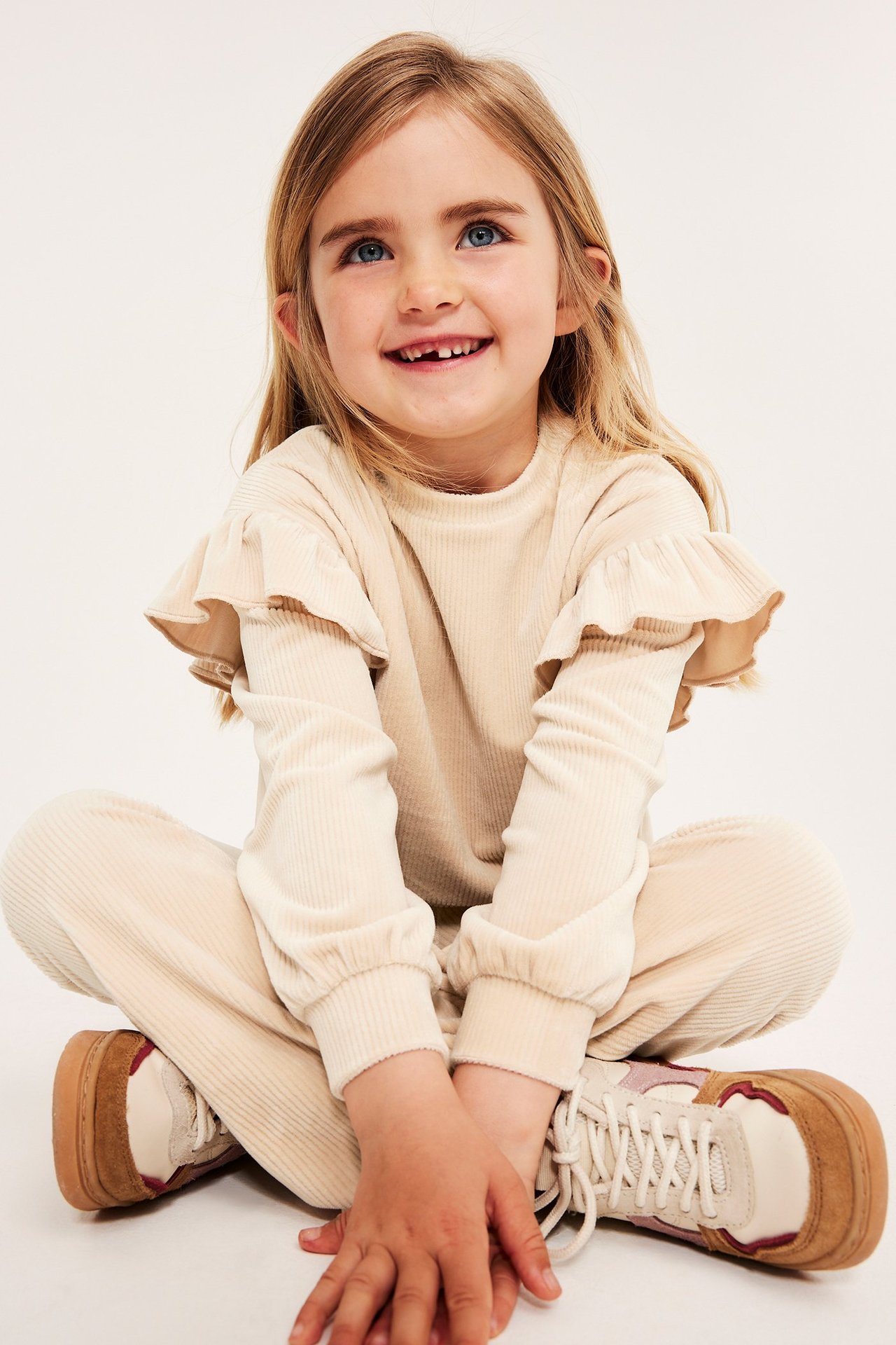 Widok z przodu: dziecko w beżowej prążkowanej welurowej bluzie z falbankami, pasujących spodniach i sneakersach.