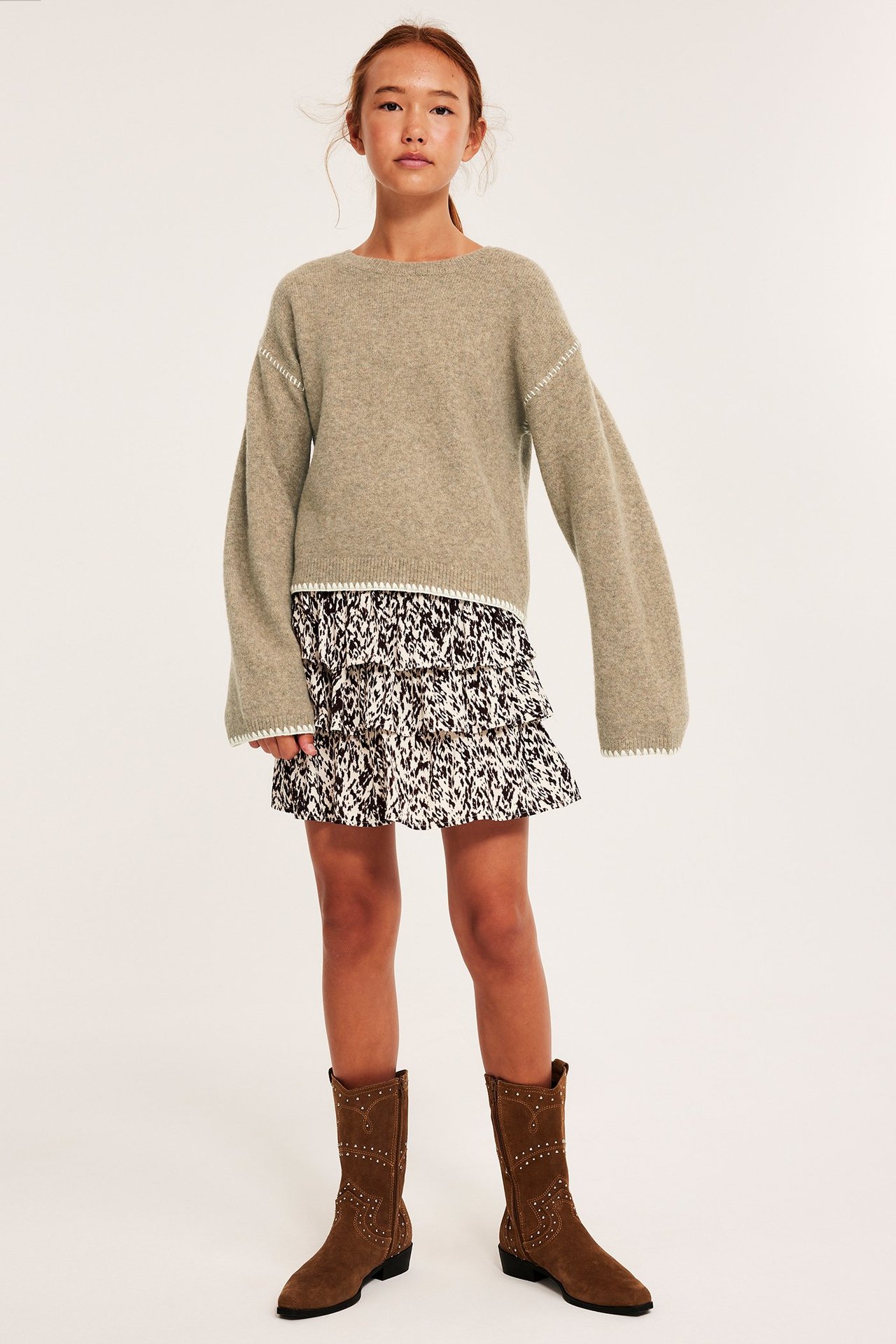 Lapsi yllään beigenvärinen neulepusero koristetikkauksilla, kuviollinen hame ja ruskeat niittisaappaat.