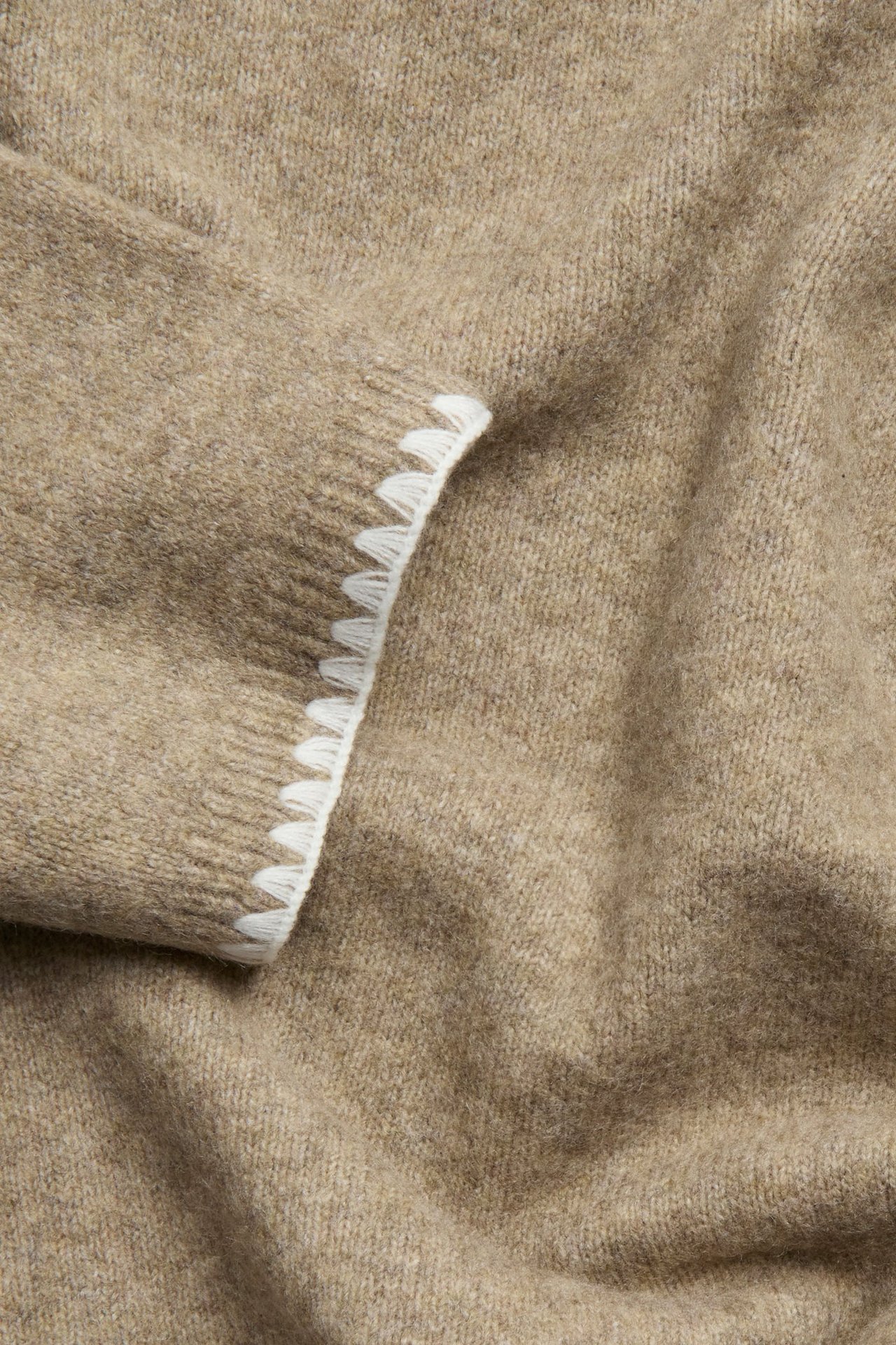Zbliżenie beżowego melanżowego swetra z białymi ozdobnymi szwami wzdłuż krawędzi.