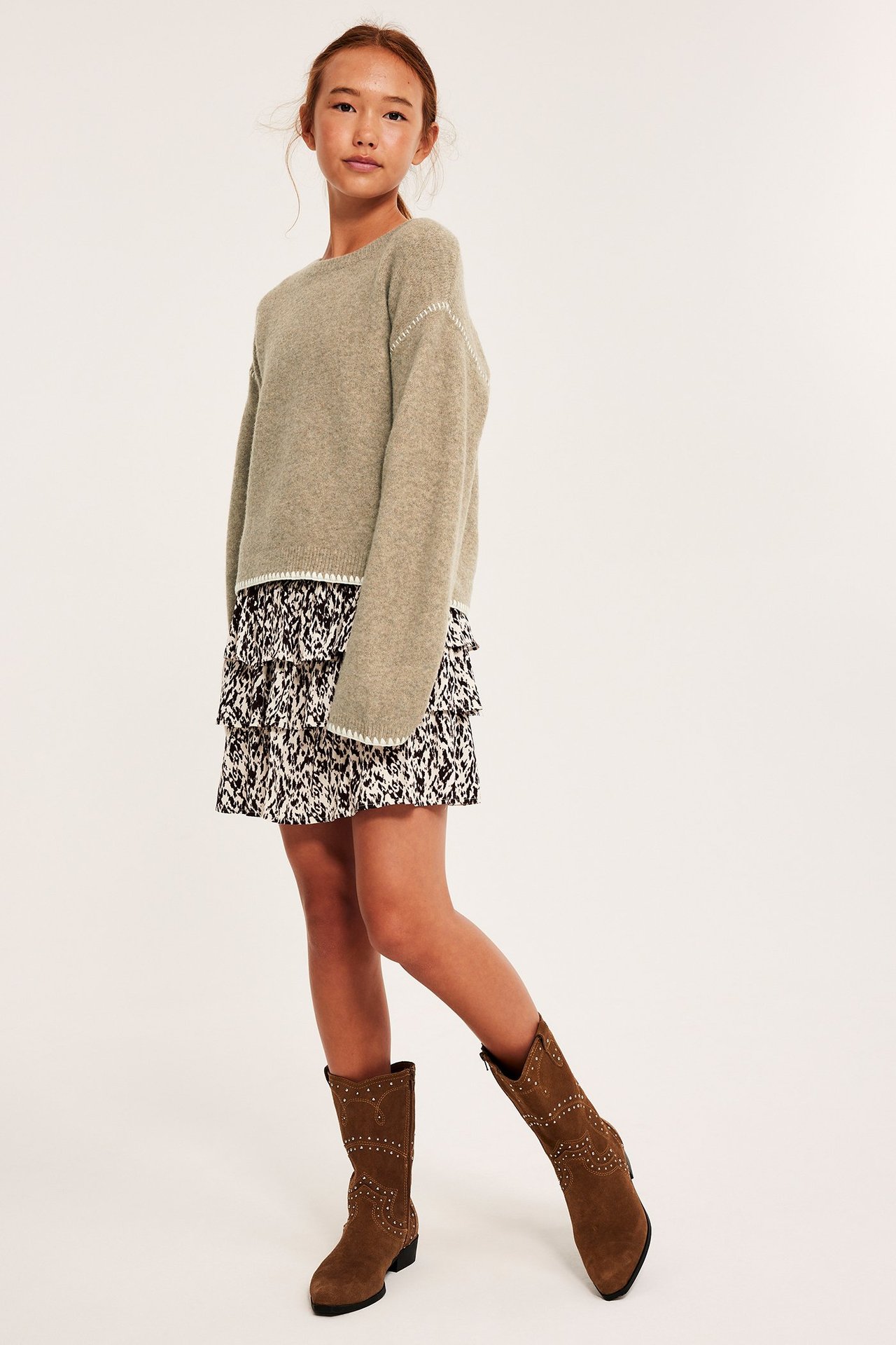 Edestäpäin lapsi beigemelange neuleessa valkoisilla tikkauksilla, kuviollisessa hameessa ja ruskeissa niittisaappaissa.