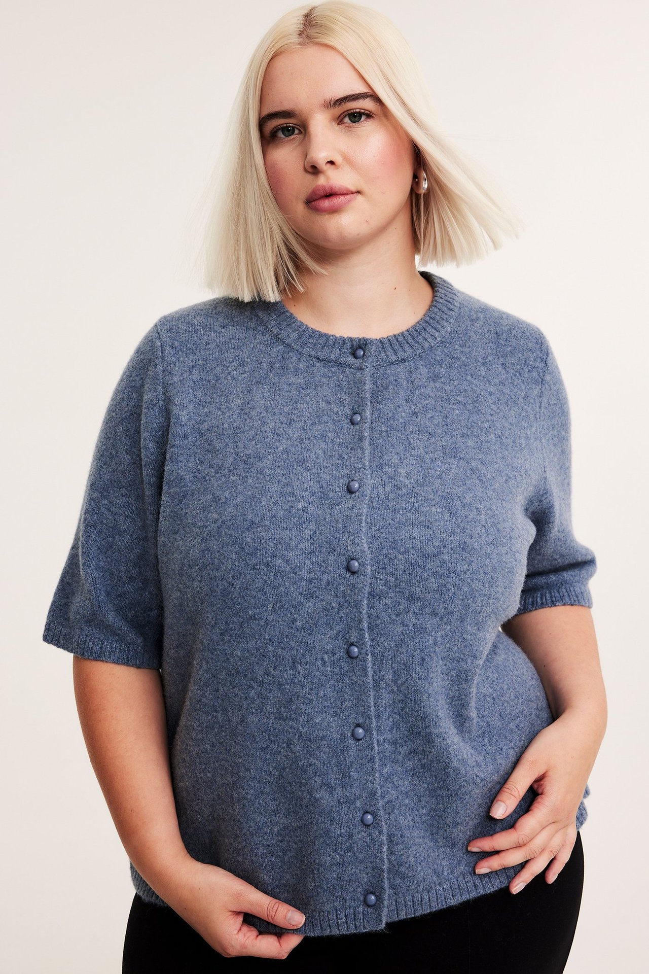 Edestä, nainen yllään sininen meleerattu lyhythihainen neuletakki napeilla mustien housujen päällä.
