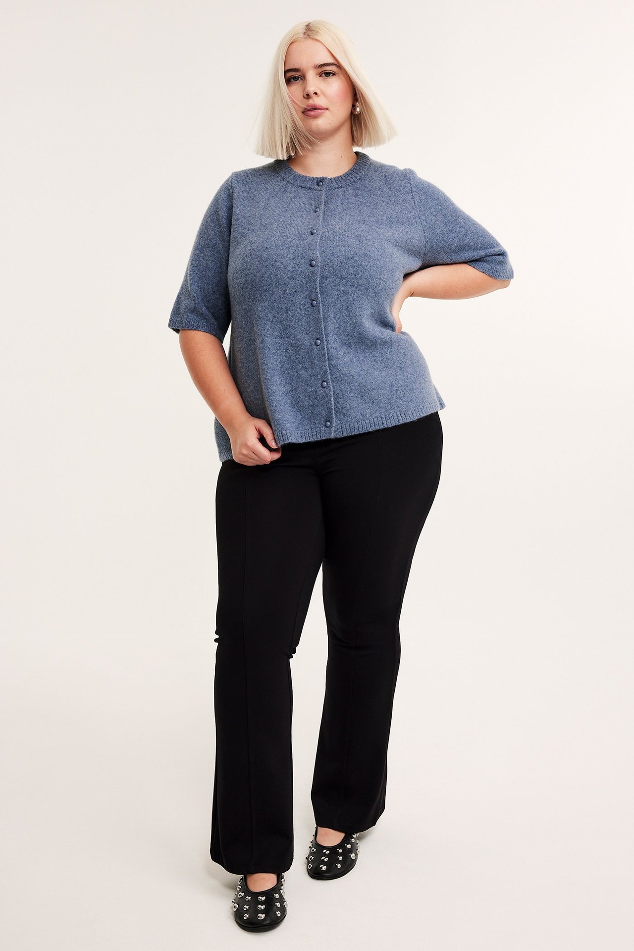 Edestäpäin: nainen yllään sininen meleerattu lyhythihainen neuletakki napeilla, mustat housut ja mustat niittikengät.