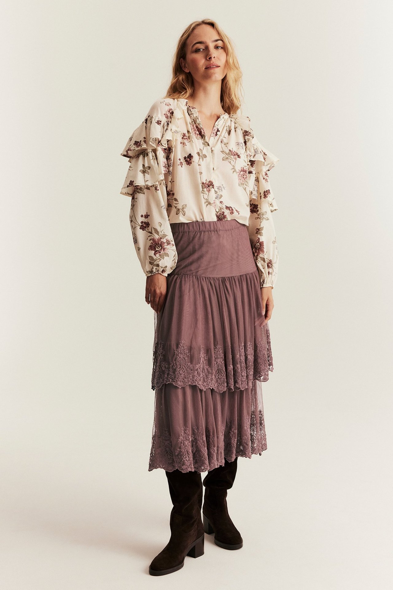 Framsida: Kvinna i mörkrosa broderad meshkjol med volanger, blommig blus och mörkbruna stövlar.