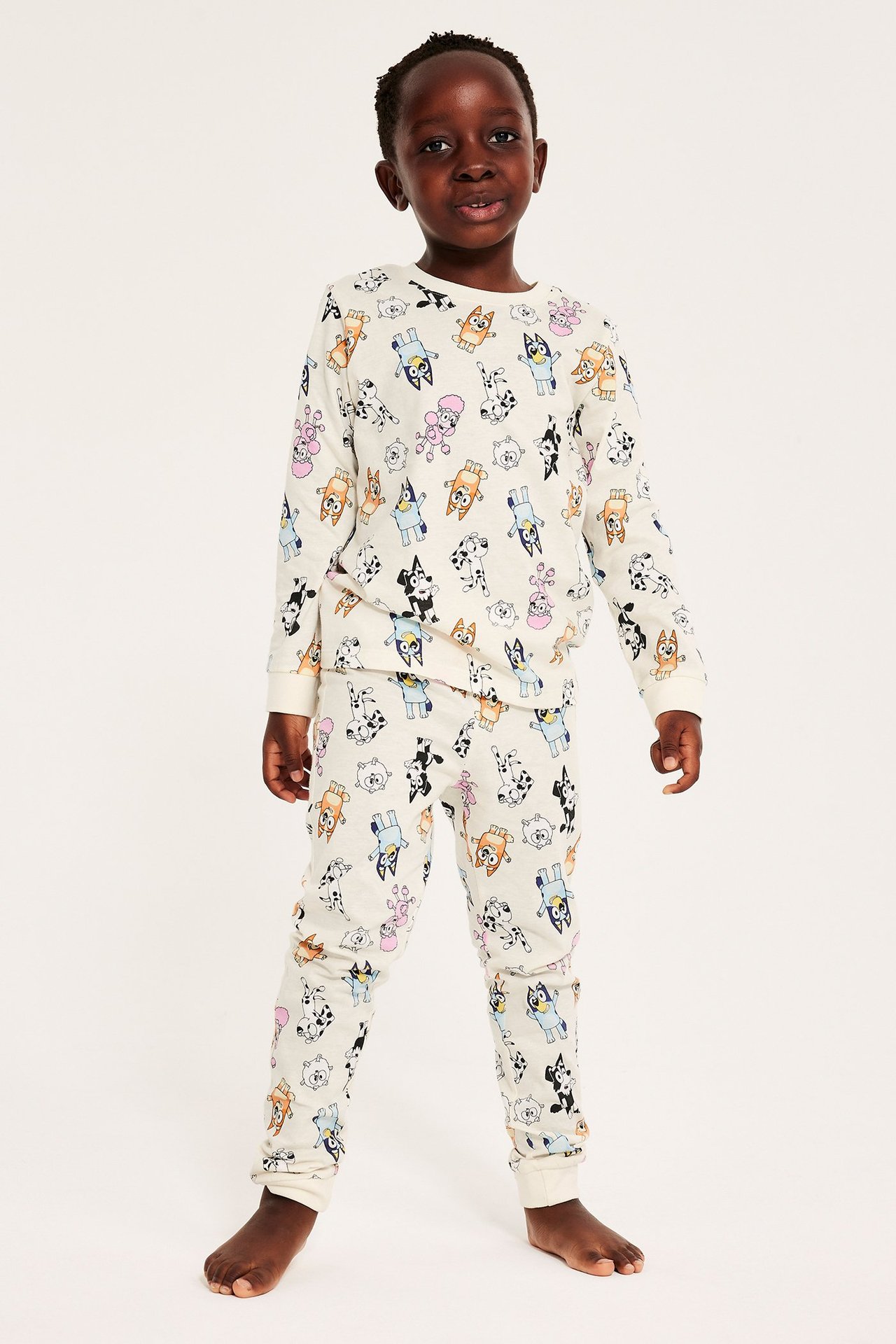 Lapsi pitkissä, kuvioiduissa pyjamissa, jossa Bluey-hahmoja, valkoinen väri.