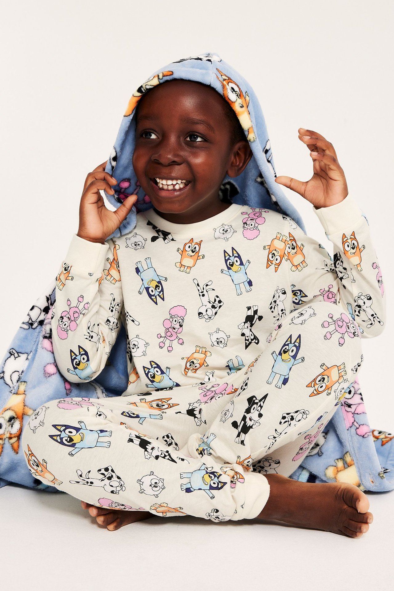 Lapsi mönstääytettyssä valko-harmaassa pyjamassa Bluey, istuu lattialla hymyillen.