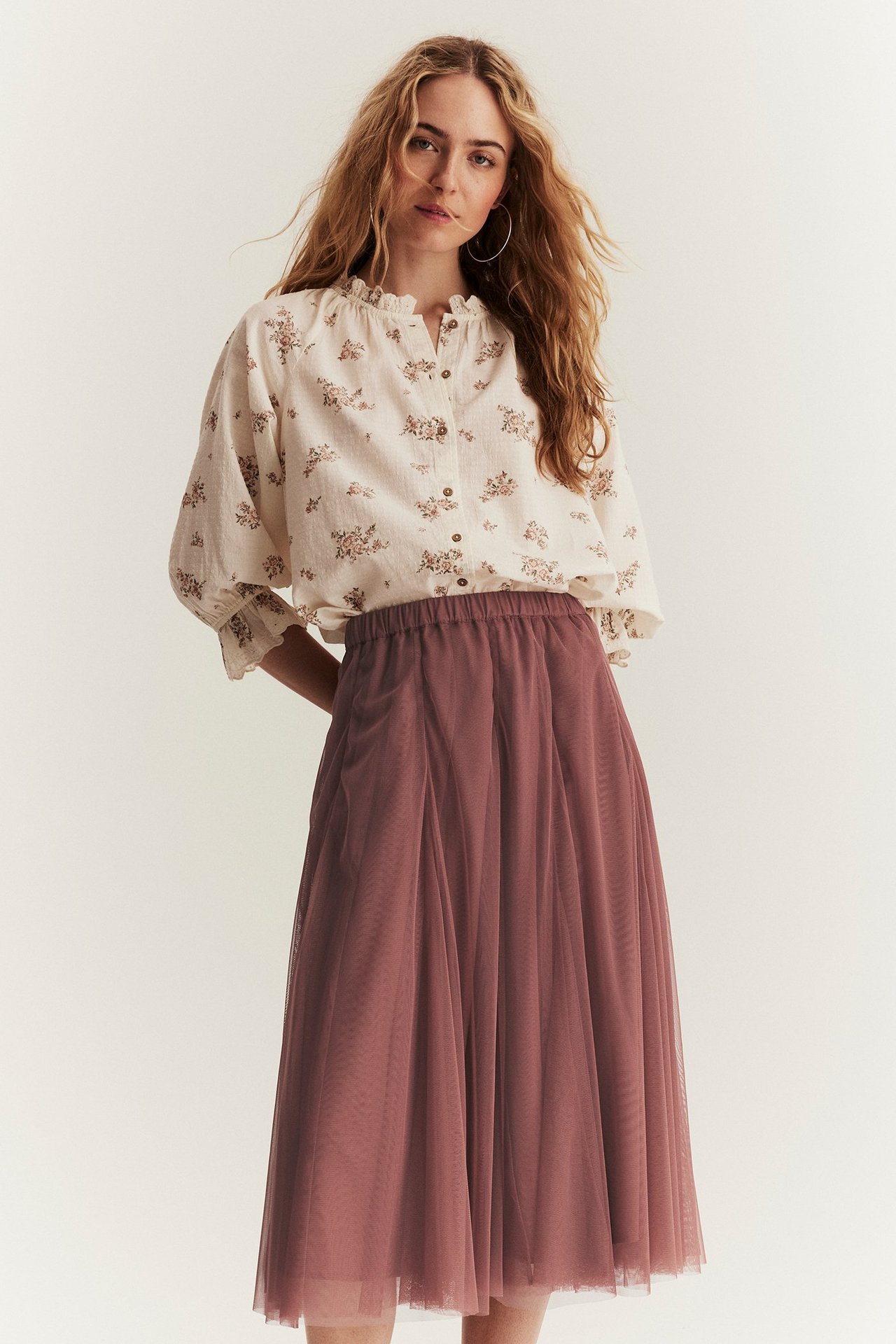 Framifrån: kvinna i mörkrosa meshkjol med resår i midjan och en krämfärgad blommig blus.