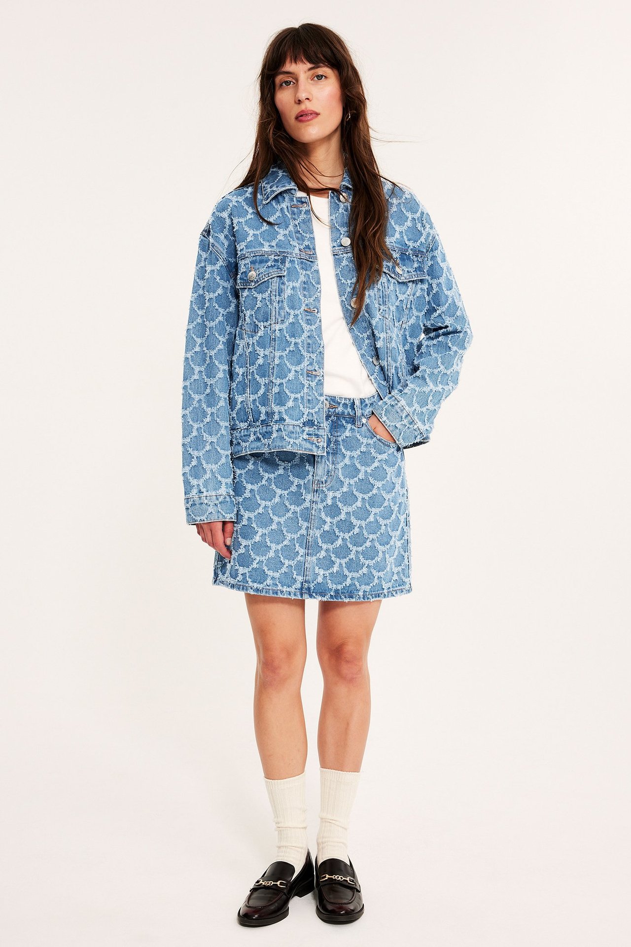 Front view: kvinne i lys blå jacquardmønstret denim miniskjørt, matchende jakke, hvit t-skjorte og svarte loafers.