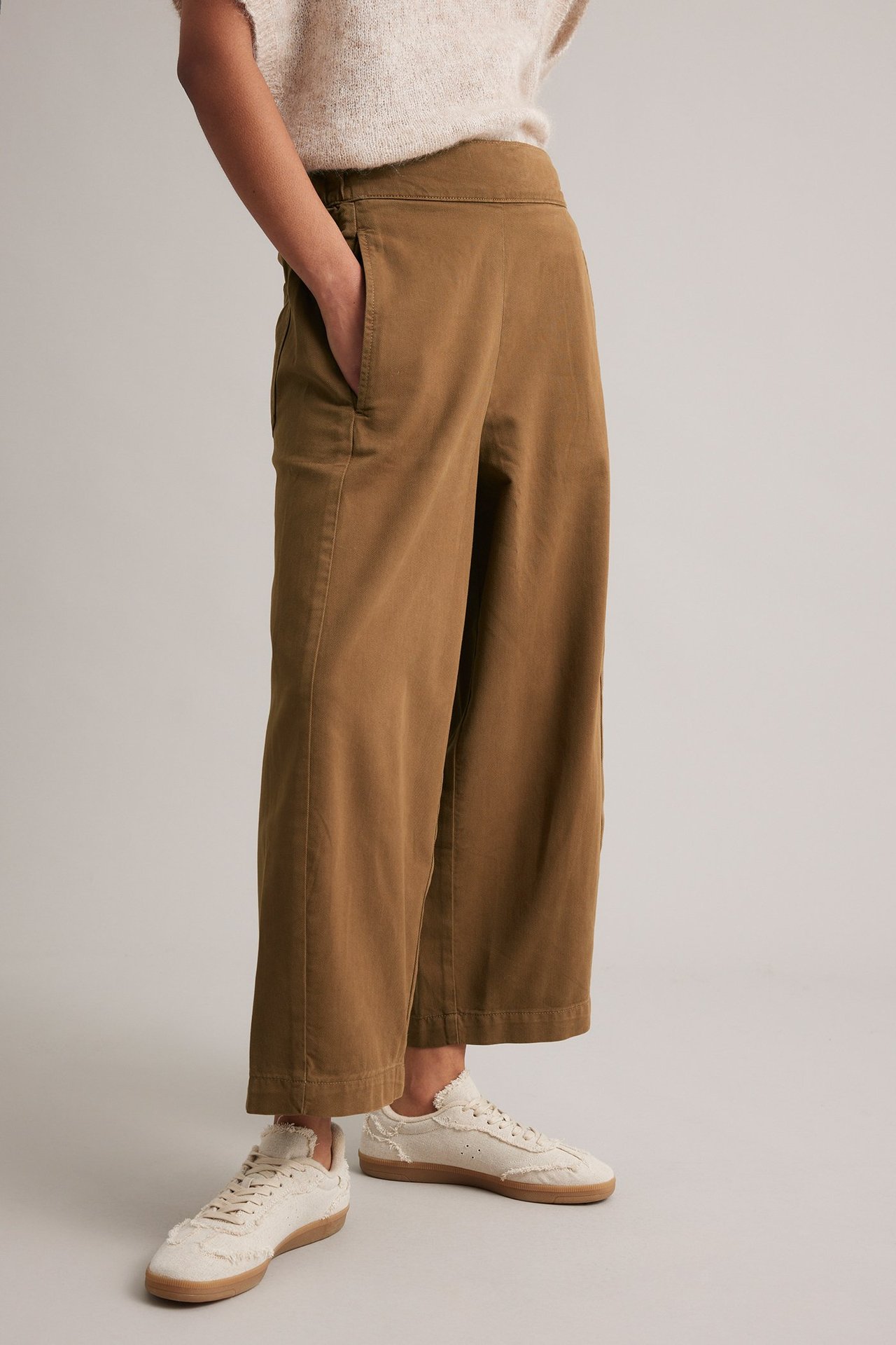 Frontvy av en kvinna i bruna vida pull on-byxor, en ljus stickad topp och vita sneakers.