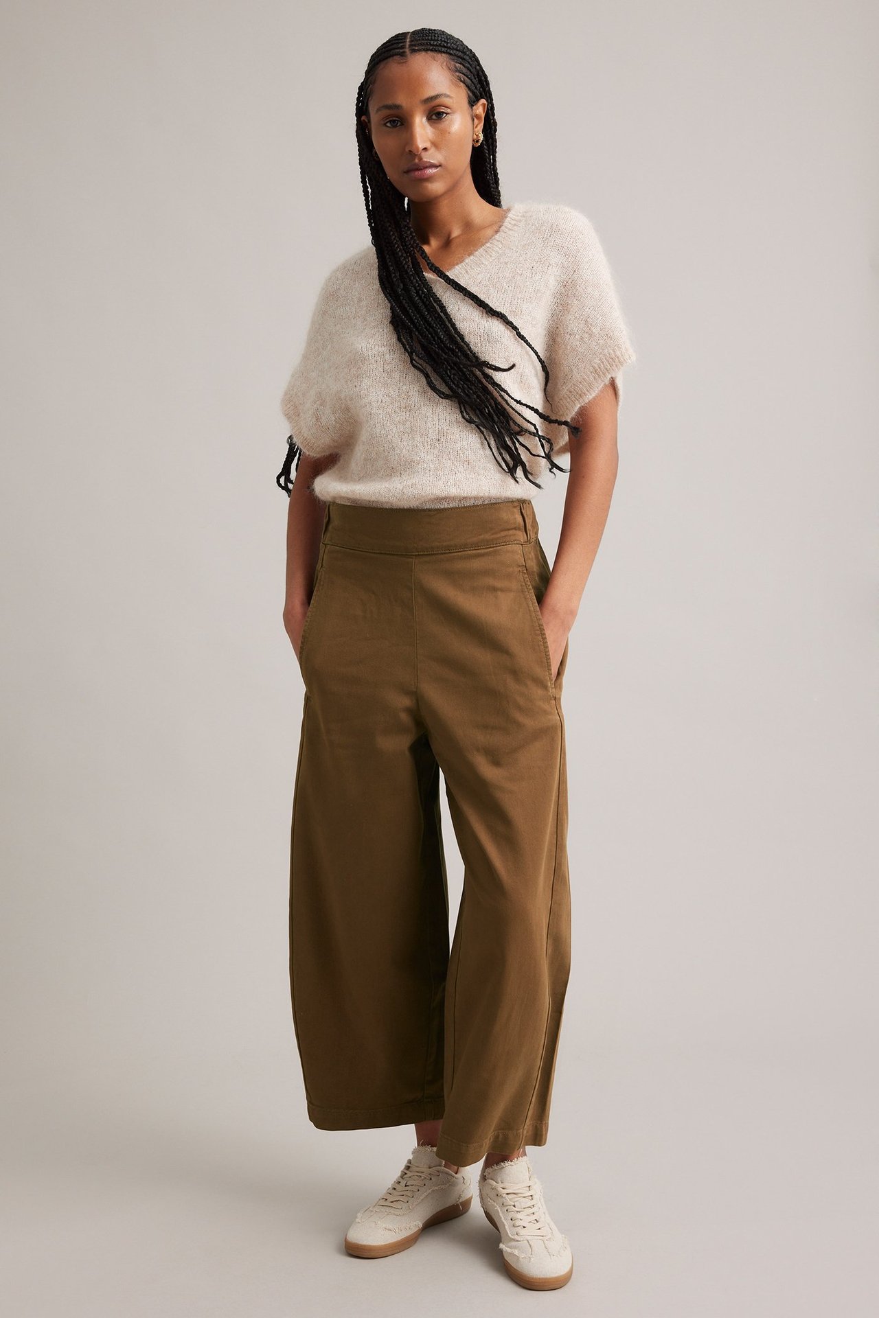 Framifrån: kvinna i bruna vida pull on-byxor, en beige kortärmad tröja och vita sneakers.