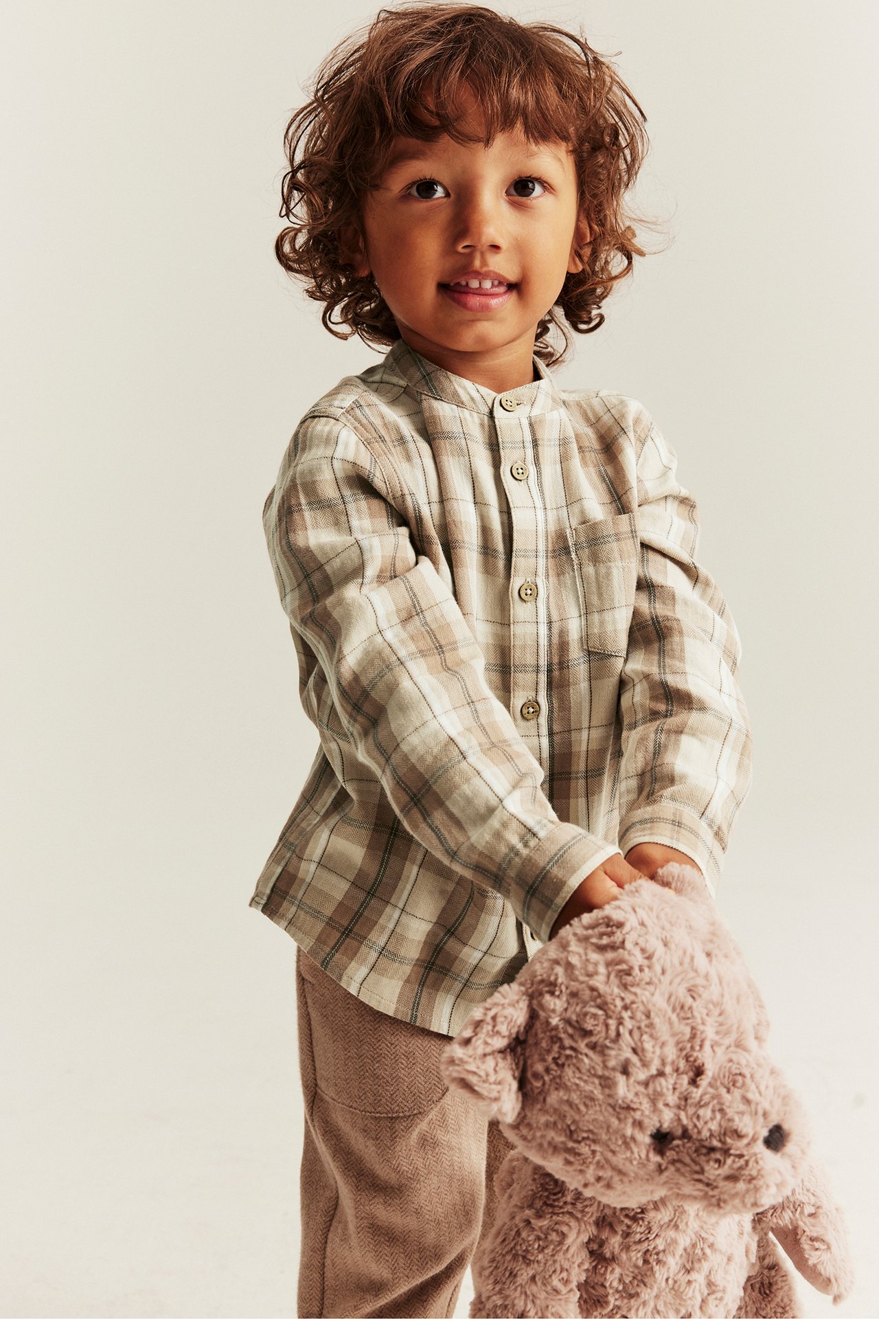 Framifrån: Barn bär beige rutig bomullsskjorta, mandarinkrage, beiga byxor, håller mjukisbjörn.
