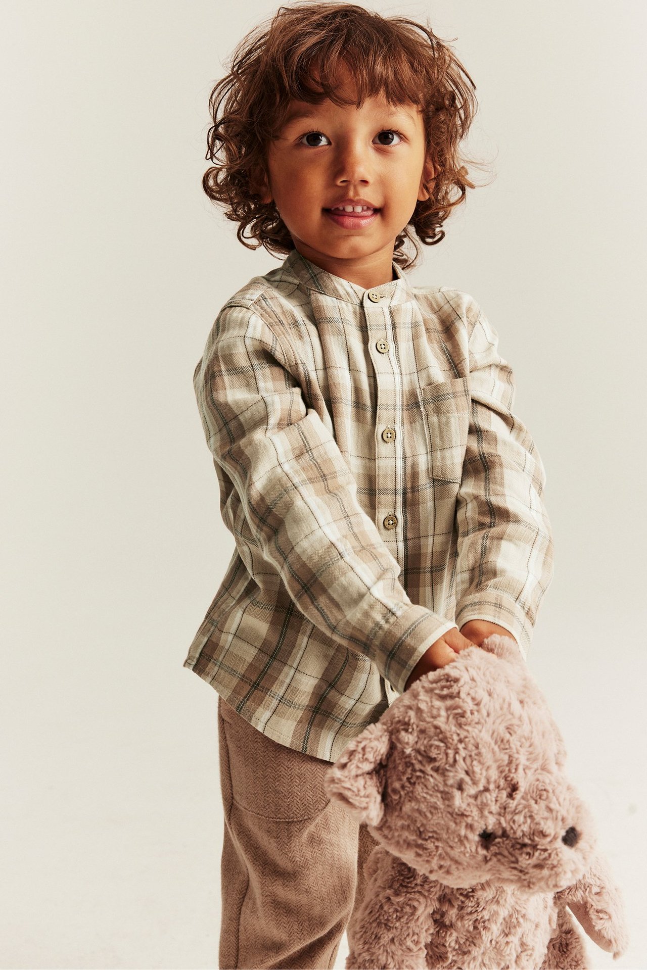 Edestä: Lapsi beigessä ruudullisessa puuvillapaidassa, mandariinikaulus, beiget housut, pitää nallea.