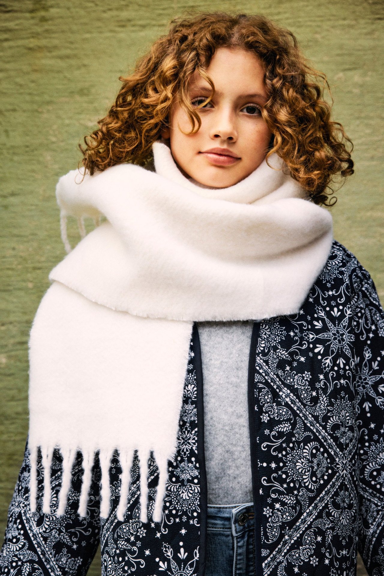 Framifrån, ett barn bär en vit fransig halsduk, en svartmönstrad jacka och en grå topp.