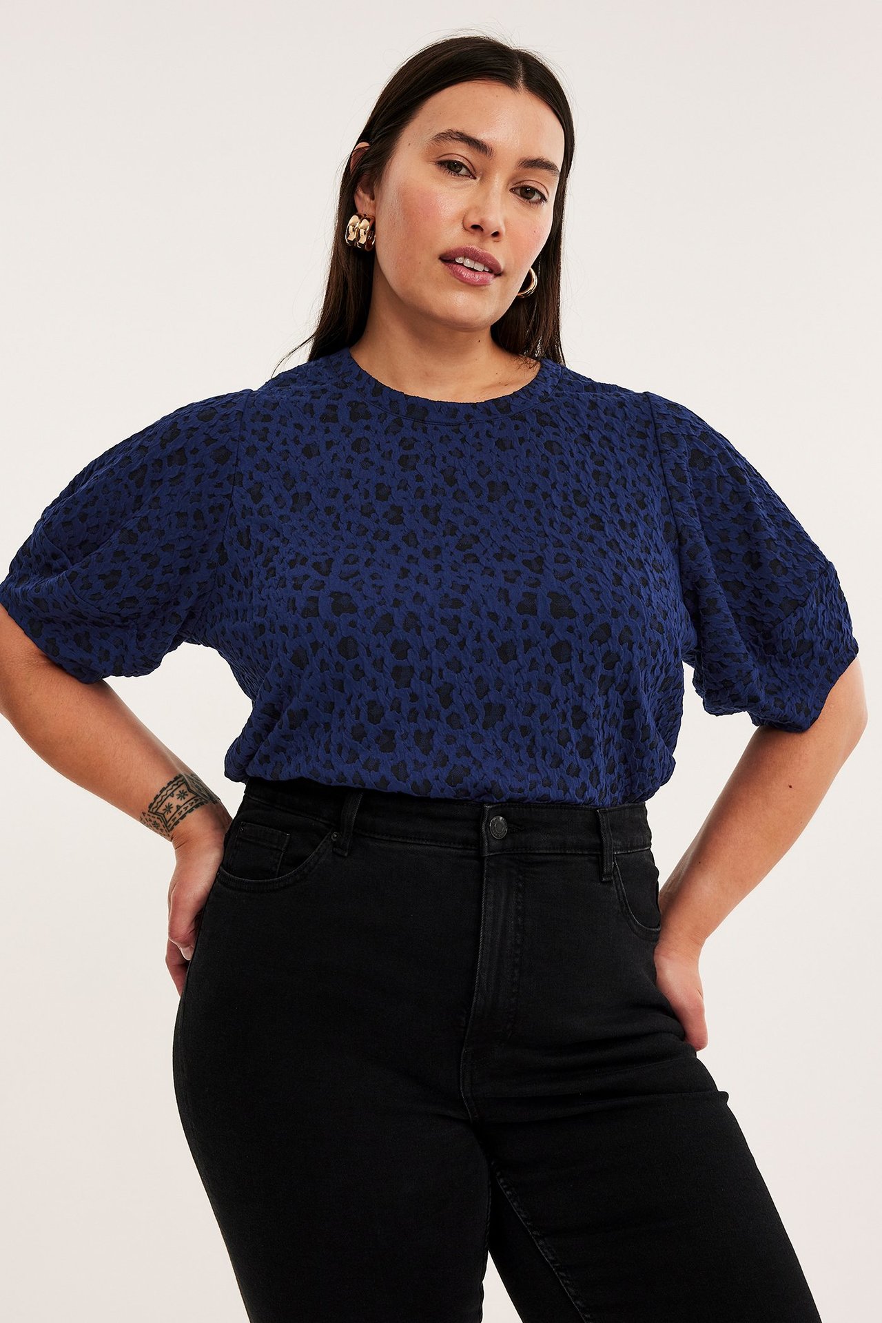 Widok z przodu: kobieta w granatowym topie w zwierzęcy wzór, czarnych jeansach i złotych kolczykach.