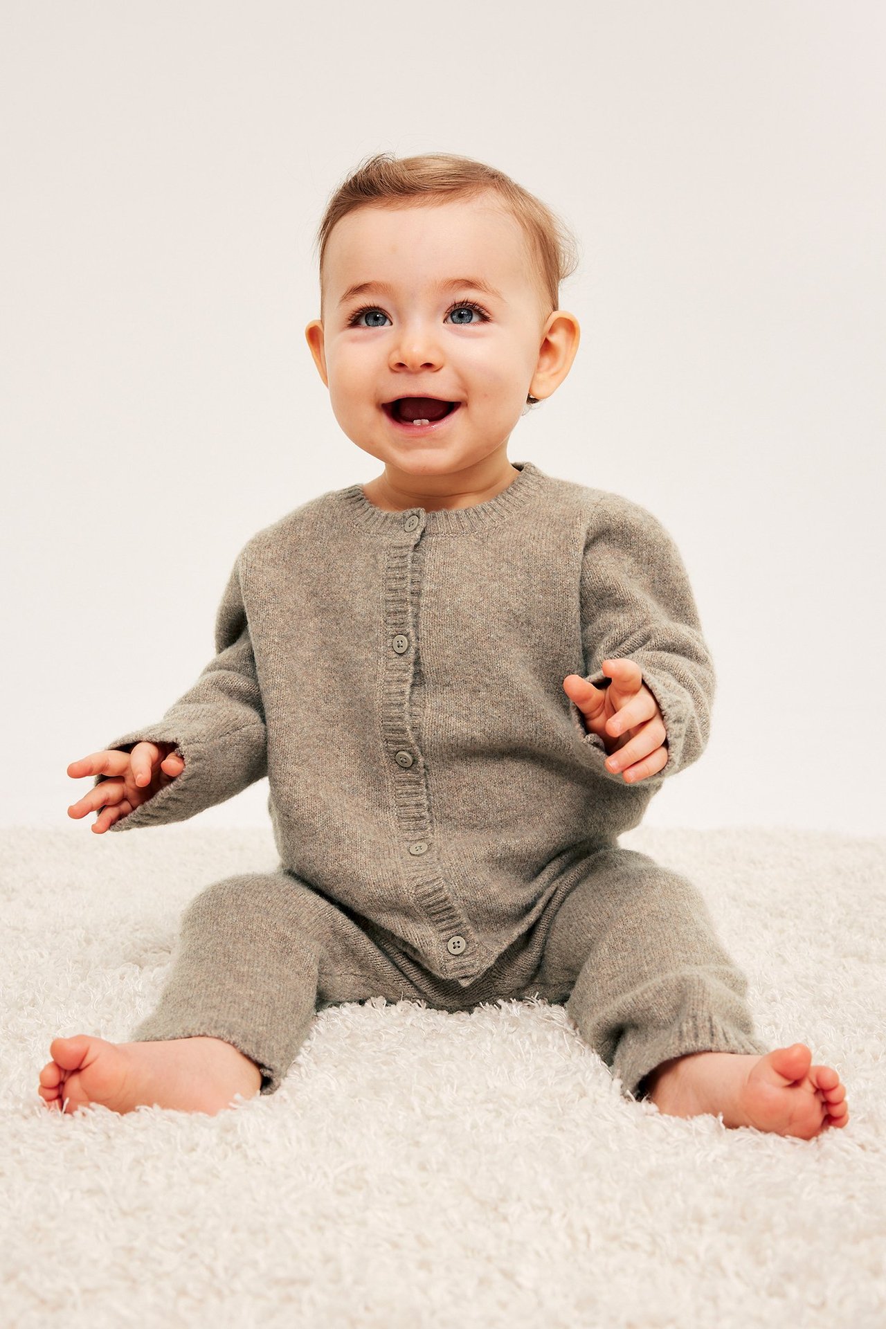 Framsida: glad baby i melerad brun stickad outfit med knappar fram, sittande på vit matta.