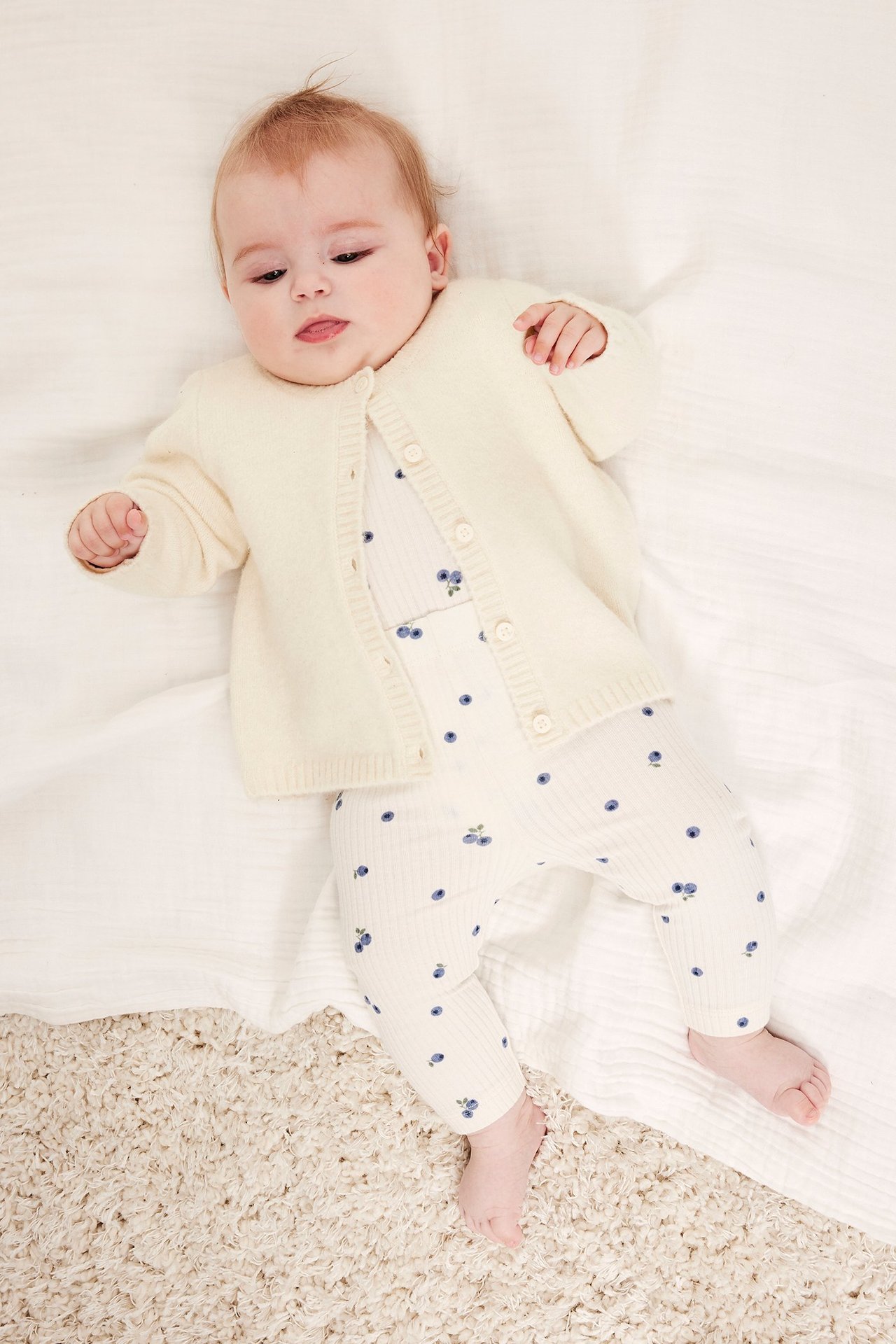 Uppifrån: baby i ljusbeige stickad kofta med knappar över vit ribbad set med blåbärstryck.