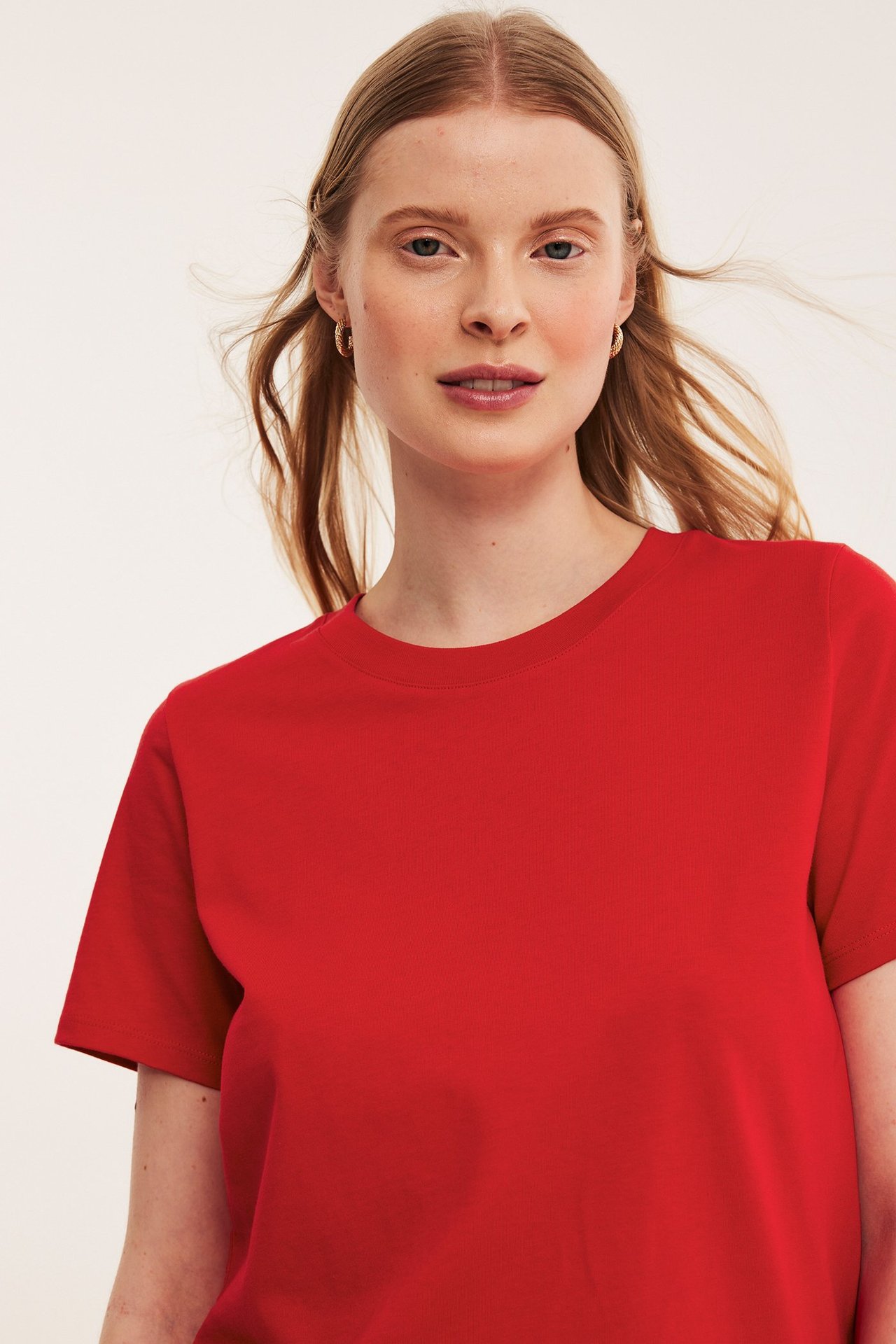Nærbilde forfra av en kvinne i en rød kortermet T-skjorte i bomull og små gulløredobber.