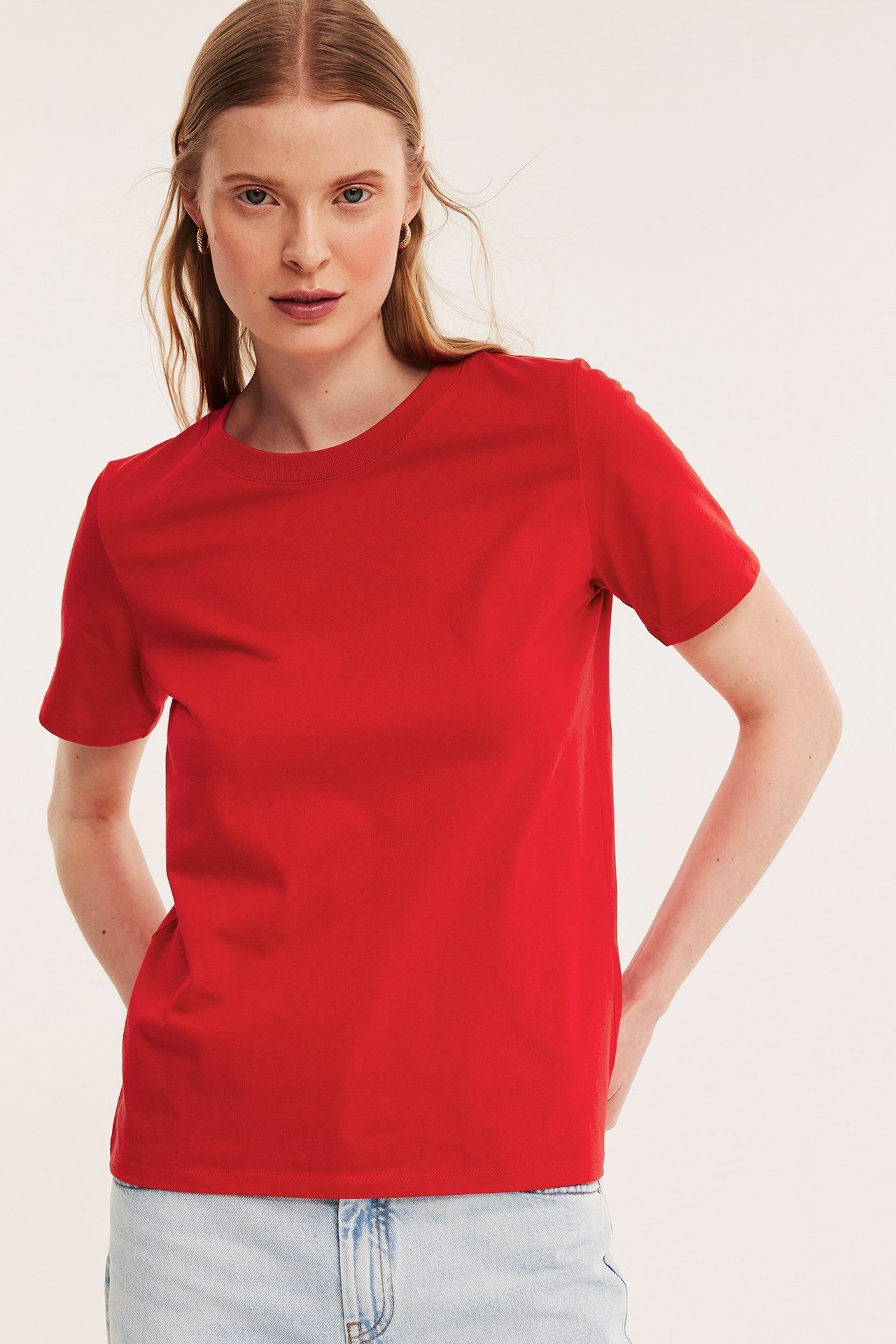 Forfra: kvinne i rød, rett passform, kortermet T-skjorte i bomull og lyseblå jeans.