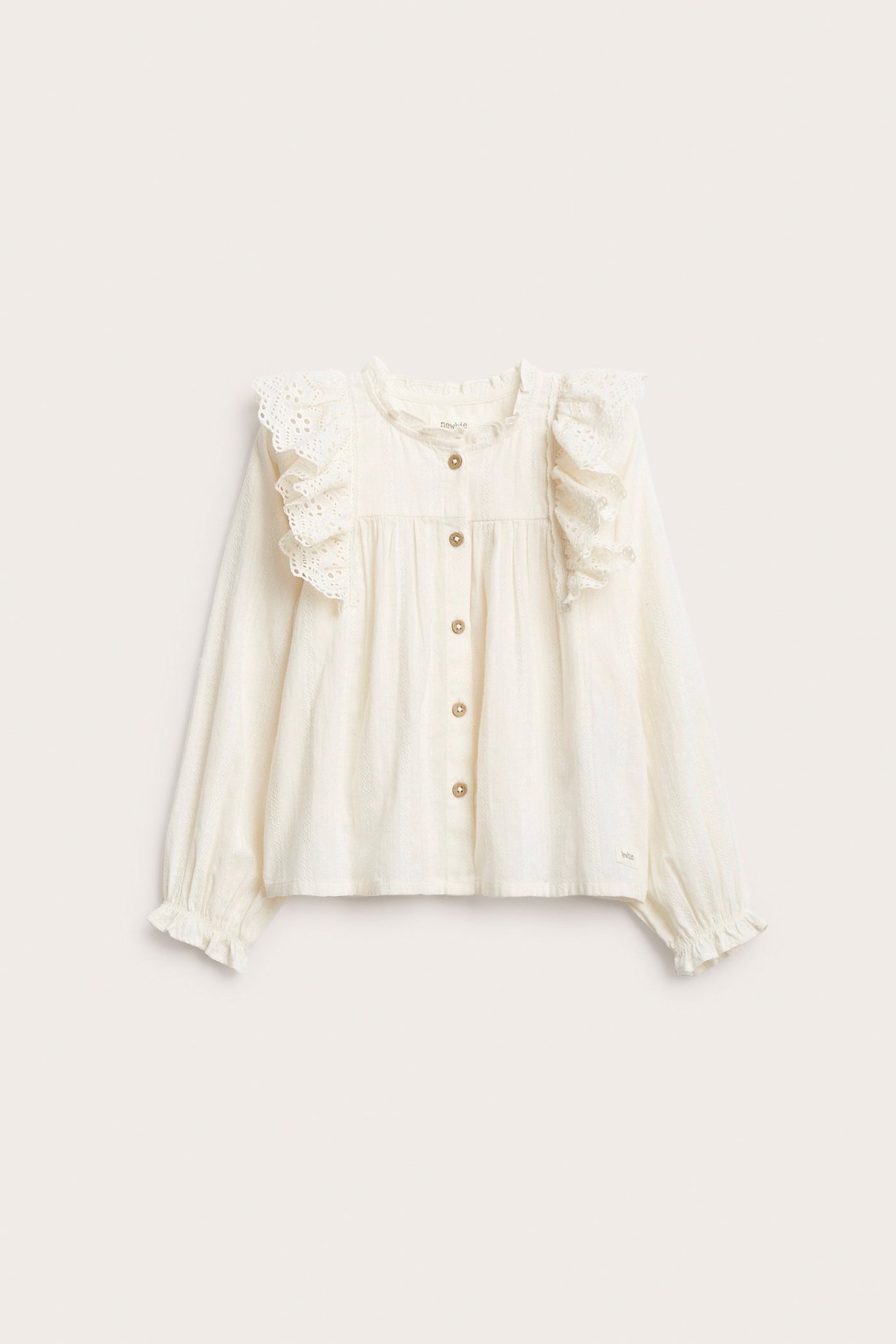 Frontvy av en offwhite barnblus i bomull med broderade volanger på axlarna och knappar framtill.