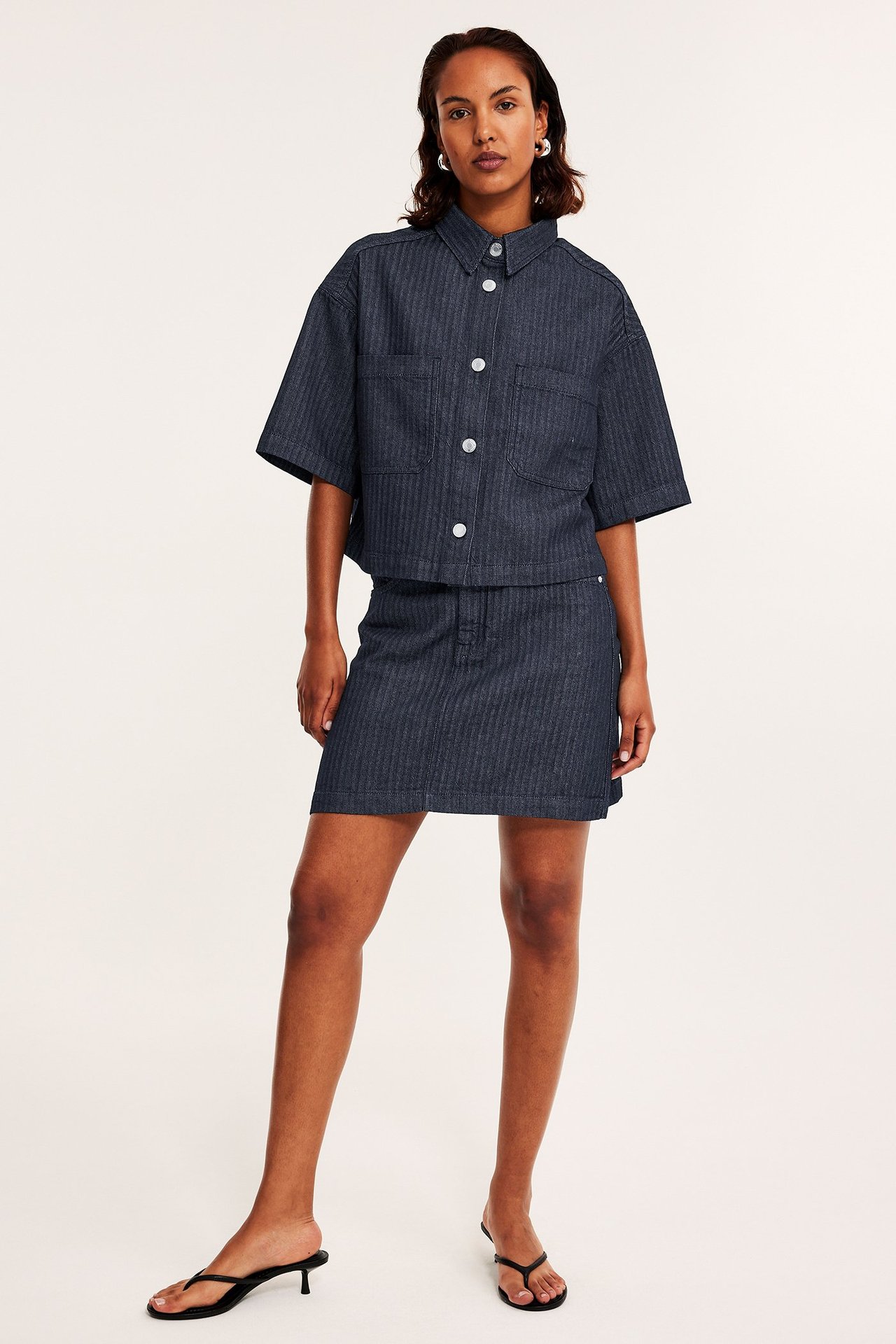 Frontvisning: Kvinne i mørk denim fiskebensmønstret miniskjørt, matchende kortermet skjorte og svarte sandaler.