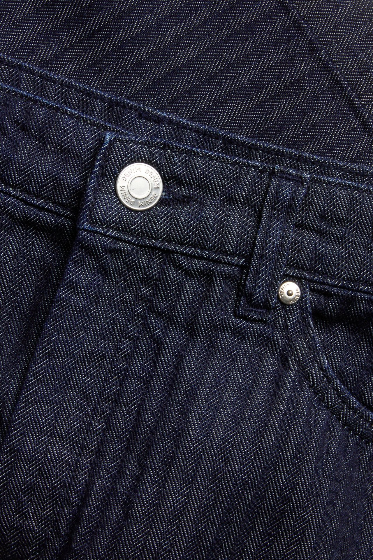 Zbliżenie ciemnej dżinsowej spódnicy we wzór w jodełkę, ze srebrnym guzikiem i nitem.