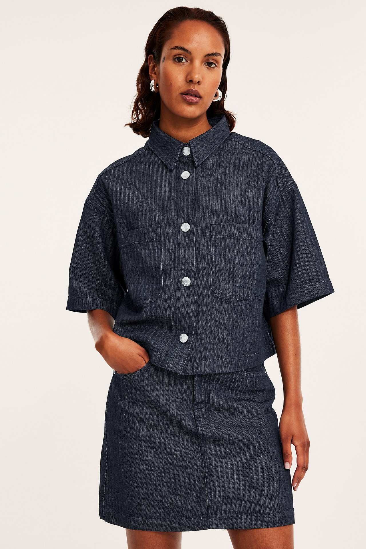 Frontvisning: Kvinne i mørk denim fiskebensmønstret kortermet skjorte med hvite knapper og matchende miniskjørt.