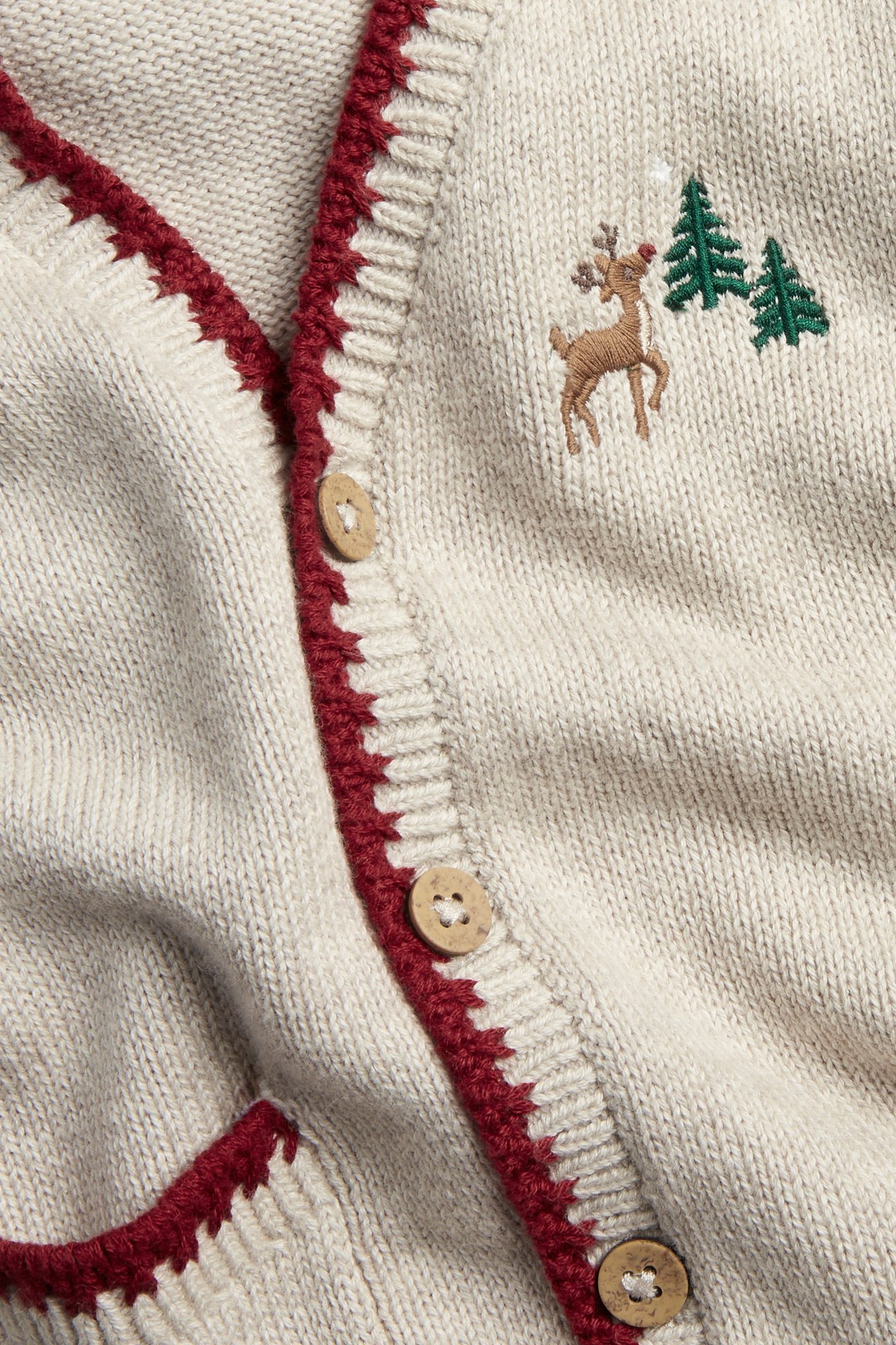 Närbild framifrån på en beige melerad stickad julväst med röd kant, broderad ren och träknappar.