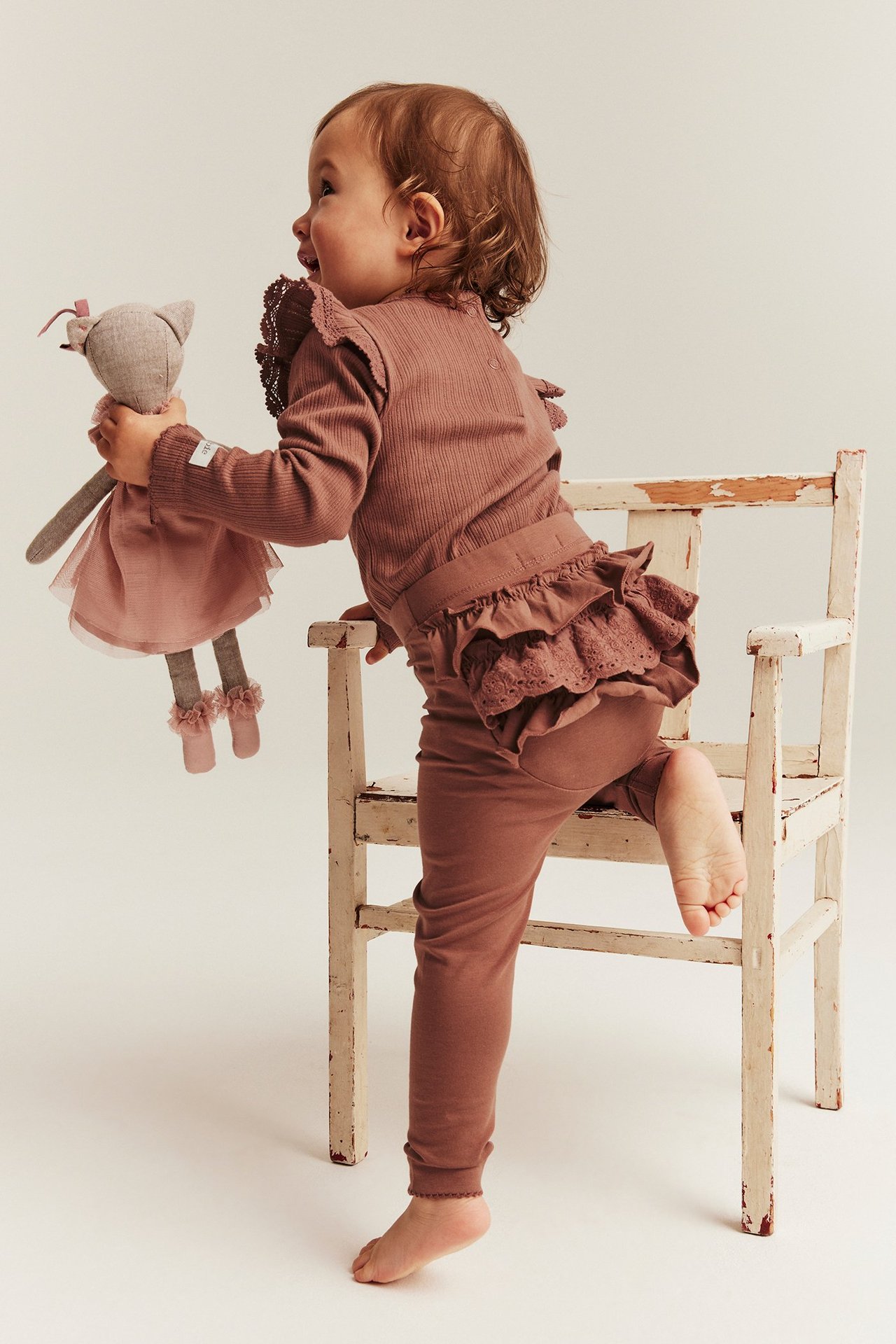 Widok z boku dziecka w różowych legginsach z falbankami i koronką, pasujący top, trzyma lalkę.
