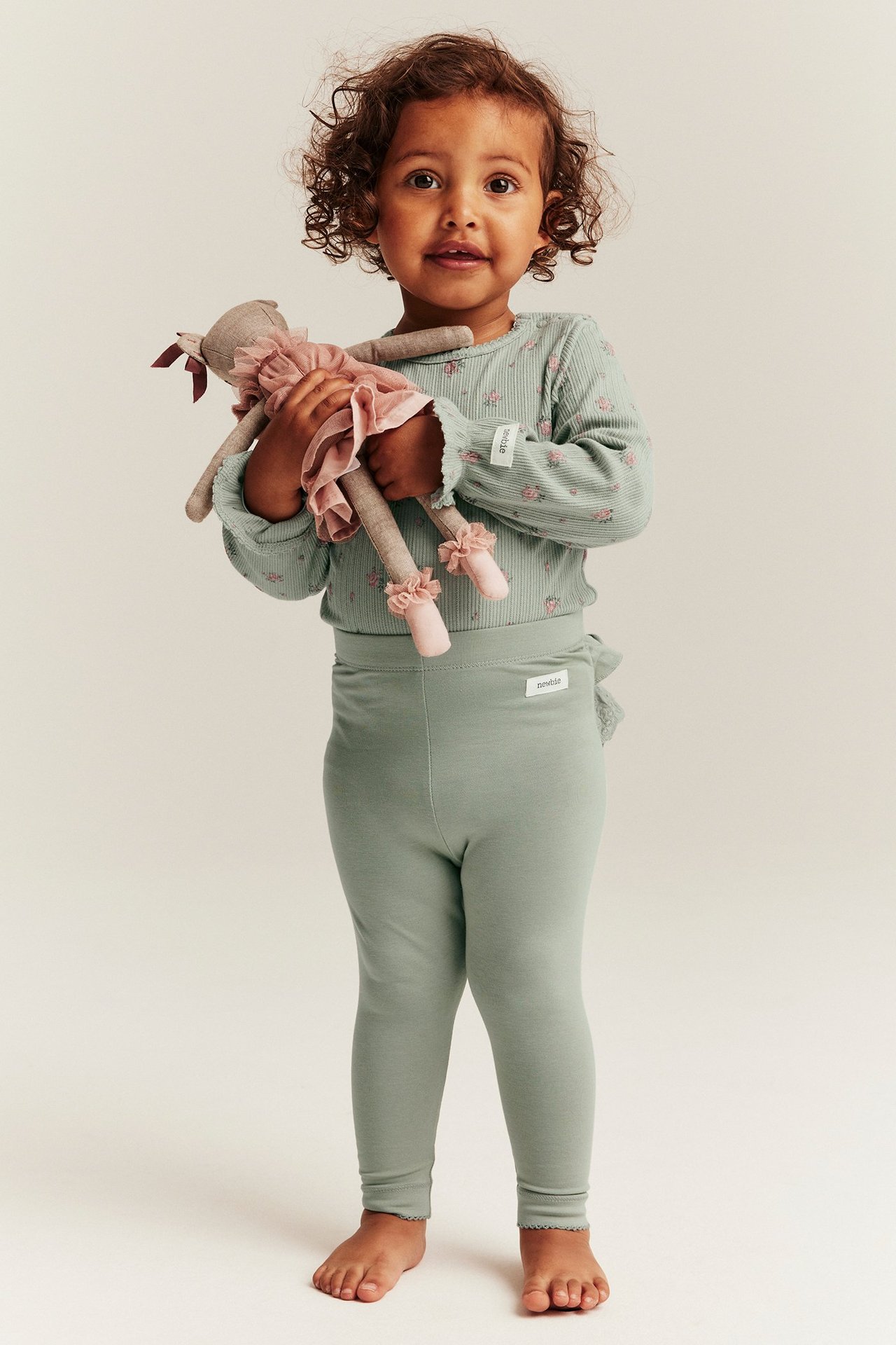 Widok z przodu dziecka w zielonych legginsach z falbankami i koronką z tyłu, oraz jasnozielonym topie, trzyma lalkę.