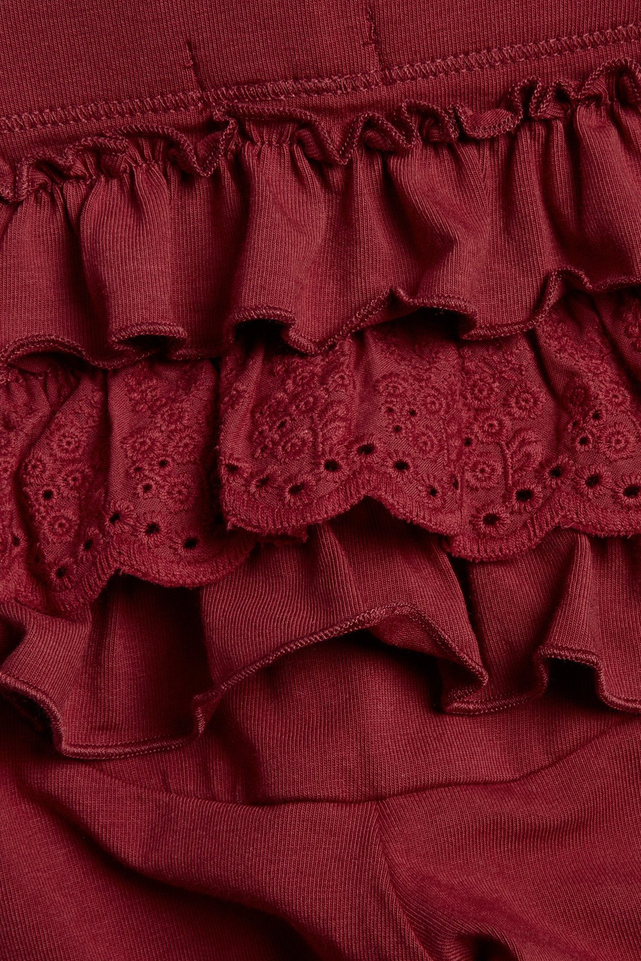 Nærbilde av mørkerøde barneleggings med elastisk midje, volanger og et lag med blonde.