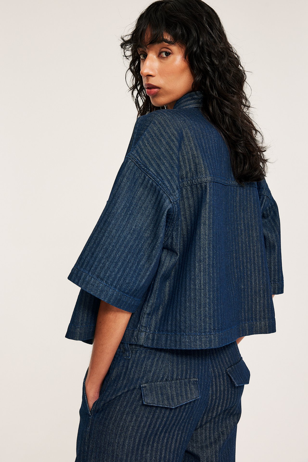 Bak-/sidebilde av en kvinne i en mørk denimstripet, kortermet crop-skjorte med matchende bukser.