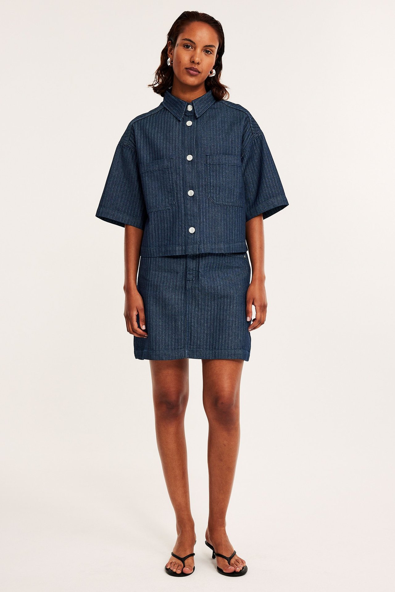 Forfra: Kvinne i mørk denim fiskebensmønstret kort skjorte, matchende miniskjørt og svarte sandaler.