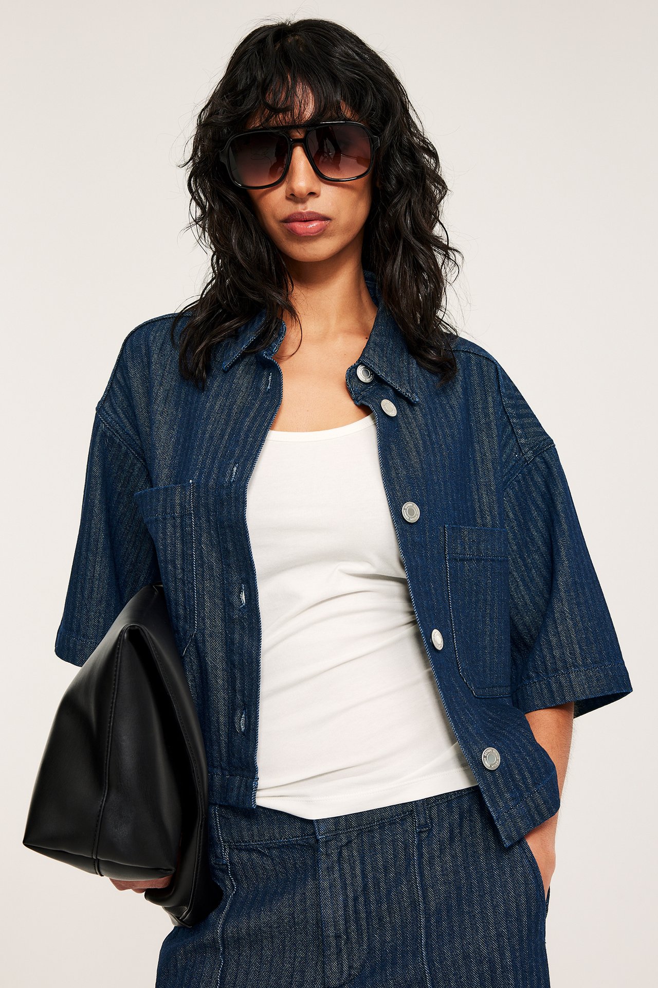 Nainen yllään tumma denim lyhyt kalanruotopaita, valkoinen toppi, housut, aurinkolasit, pitää mustaa laukkua.