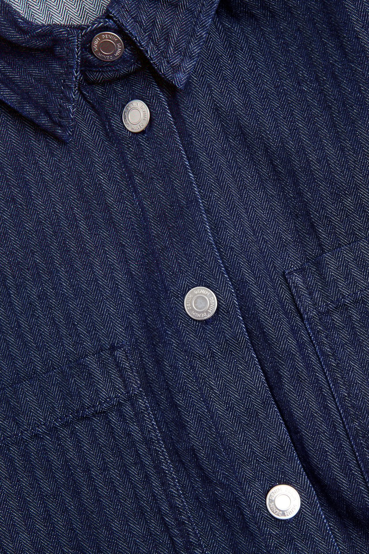 Zbliżenie na ciemnoniebieską koszulę jeansową z wzorem w jodełkę i srebrnymi guzikami.