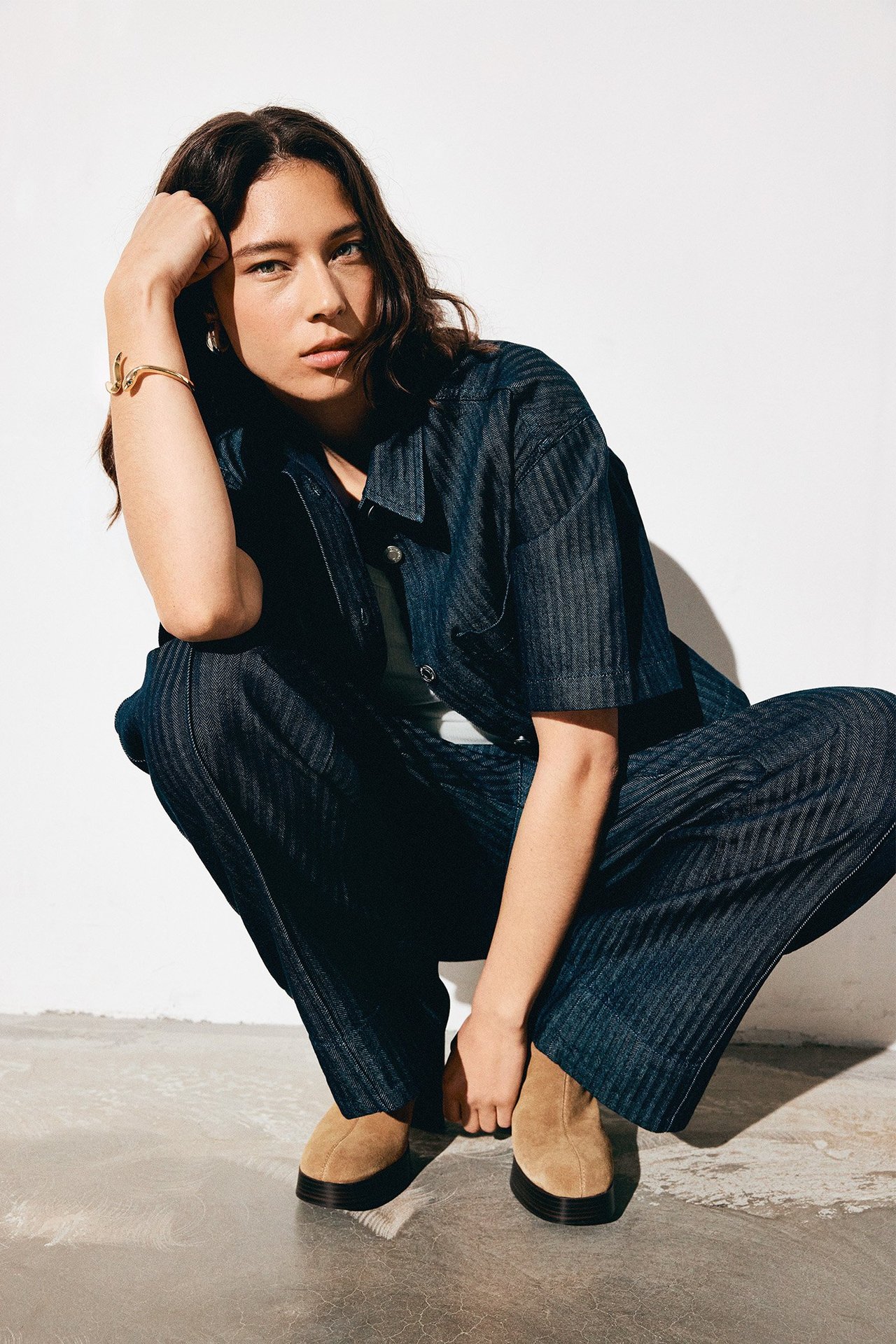 Front: Kvinne i mørk denim fiskebensmønstret kort skjorte, matchende bukser, lys topp, beige sko, gullarmbånd.