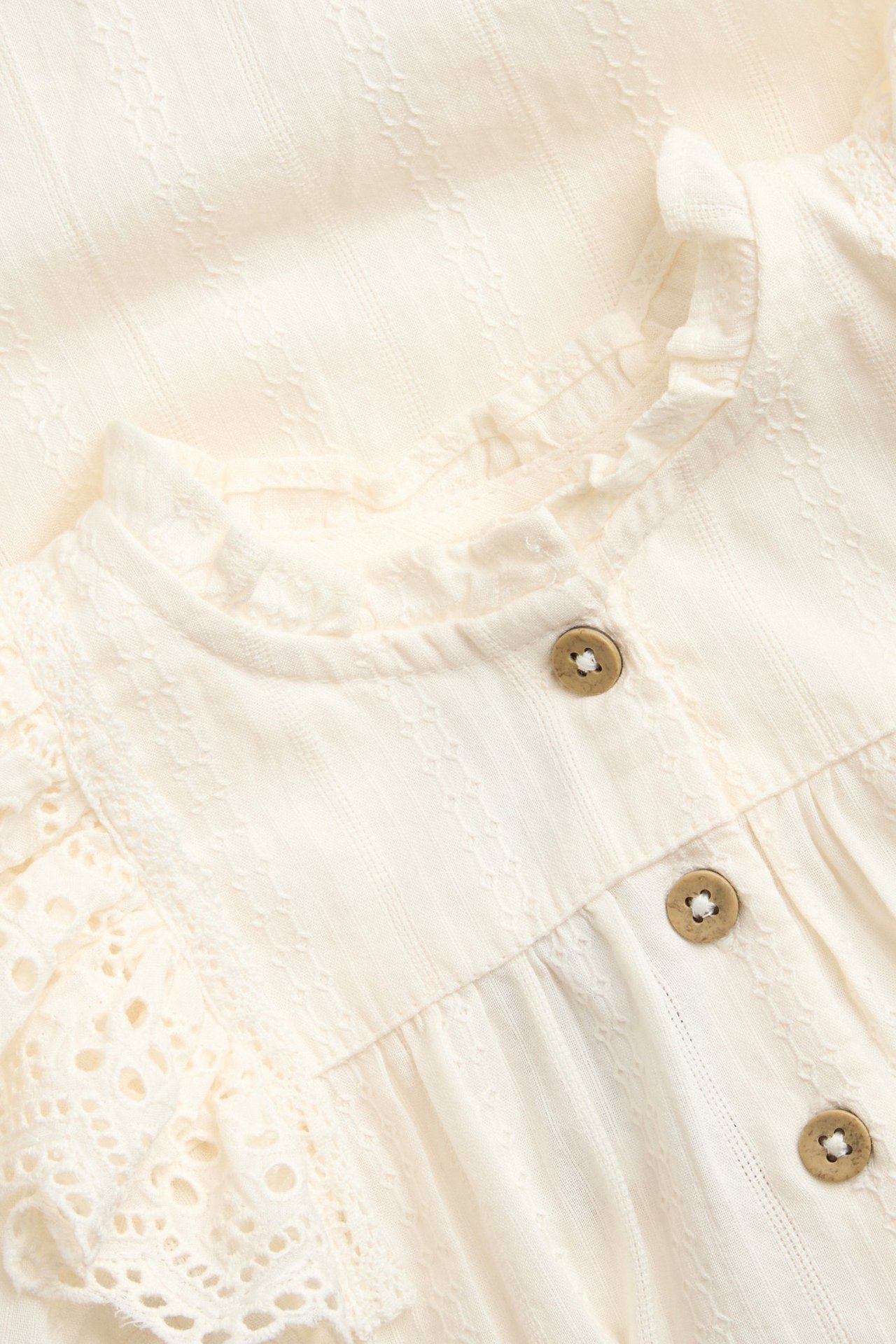 Zbliżenie, przód: ecru bawełniane body niemowlęce z tkaniny dobby, falbankami i guzikami.
